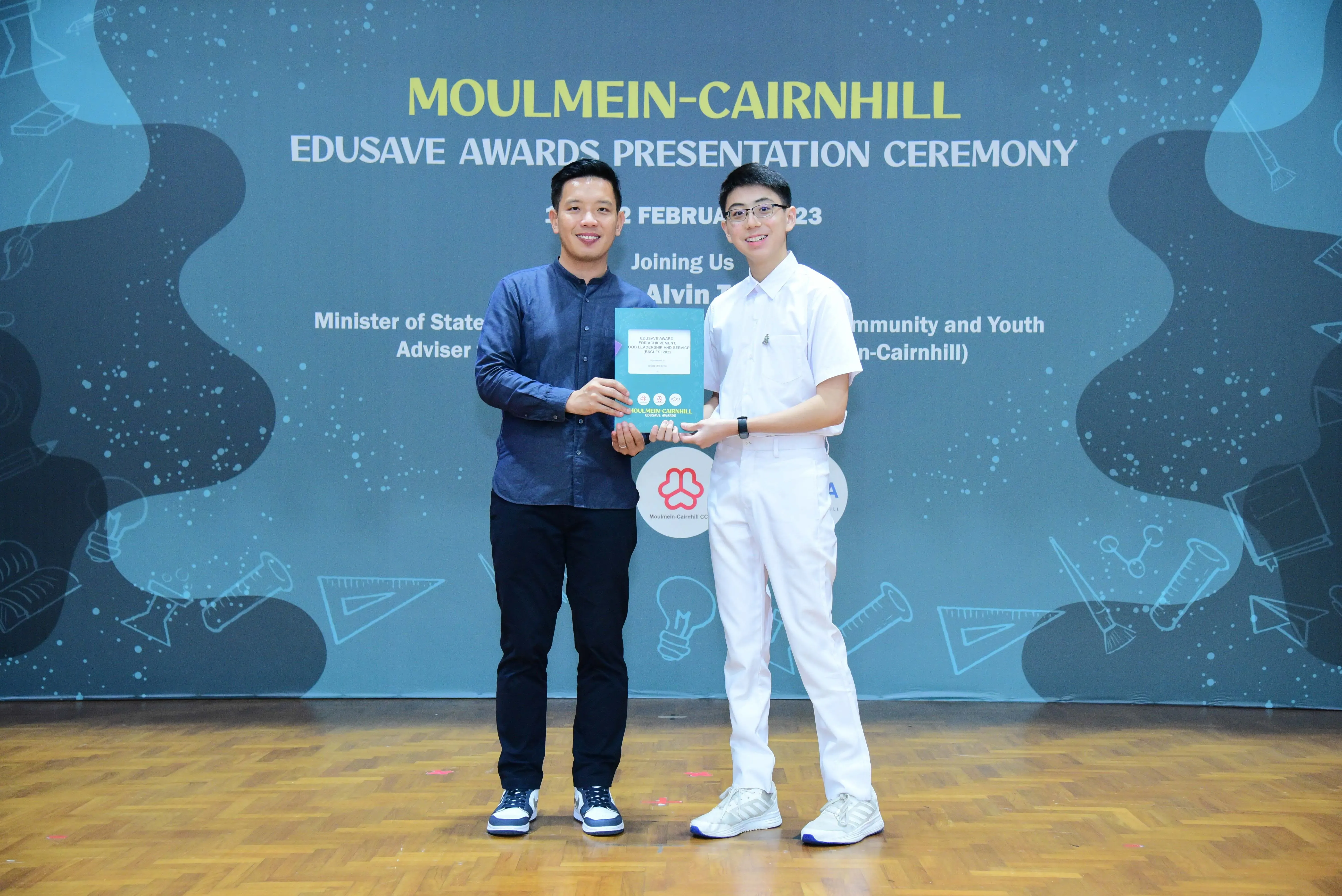 Two men smile holding an award booklet at the Moulmein-Cairnhill Edusave Awards Presentation Ceremony backdrop.