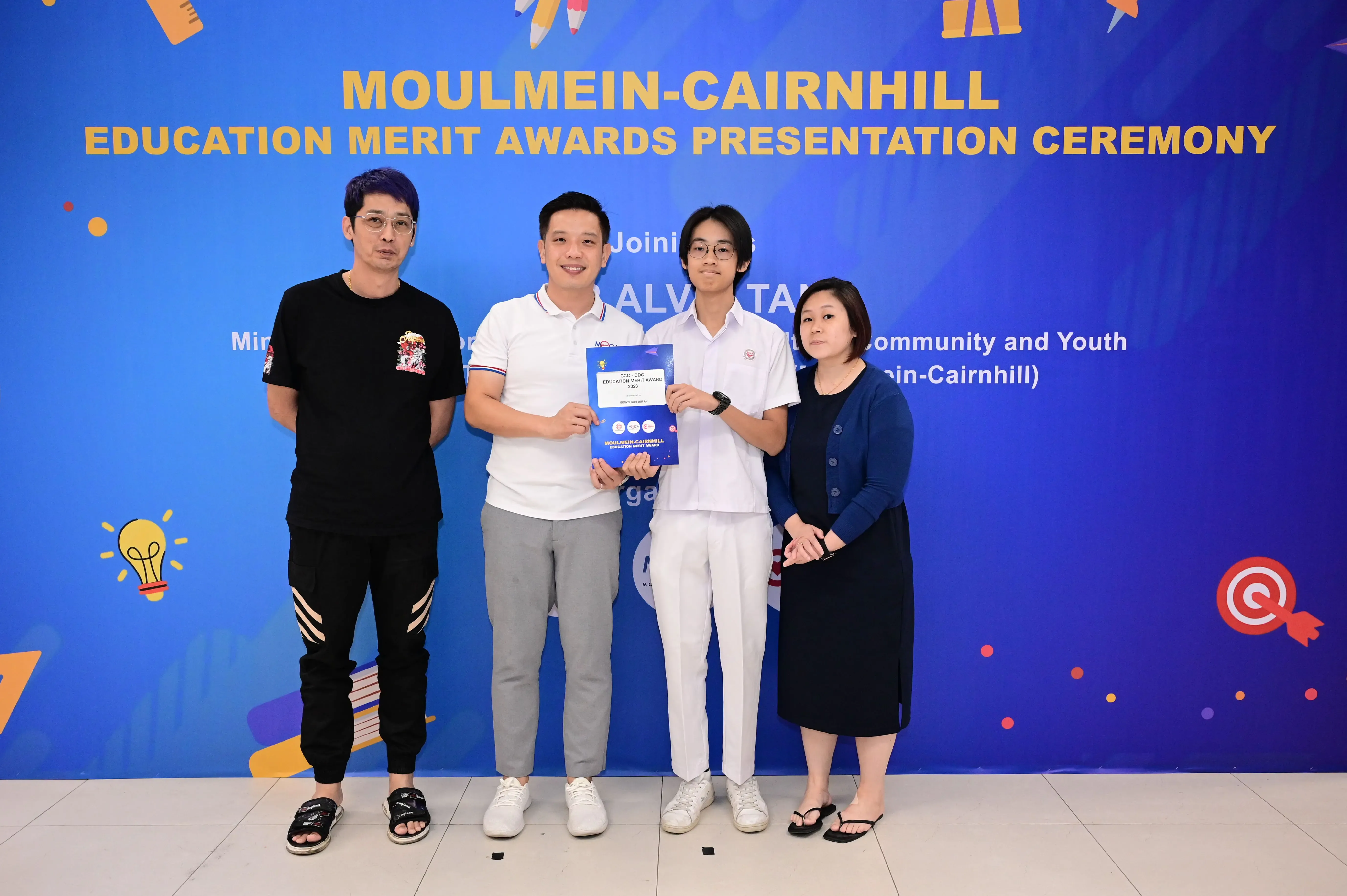 Four people stand before a blue Moulmein-Cairnhill Education Merit Awards banner, two holding a blue certificate.