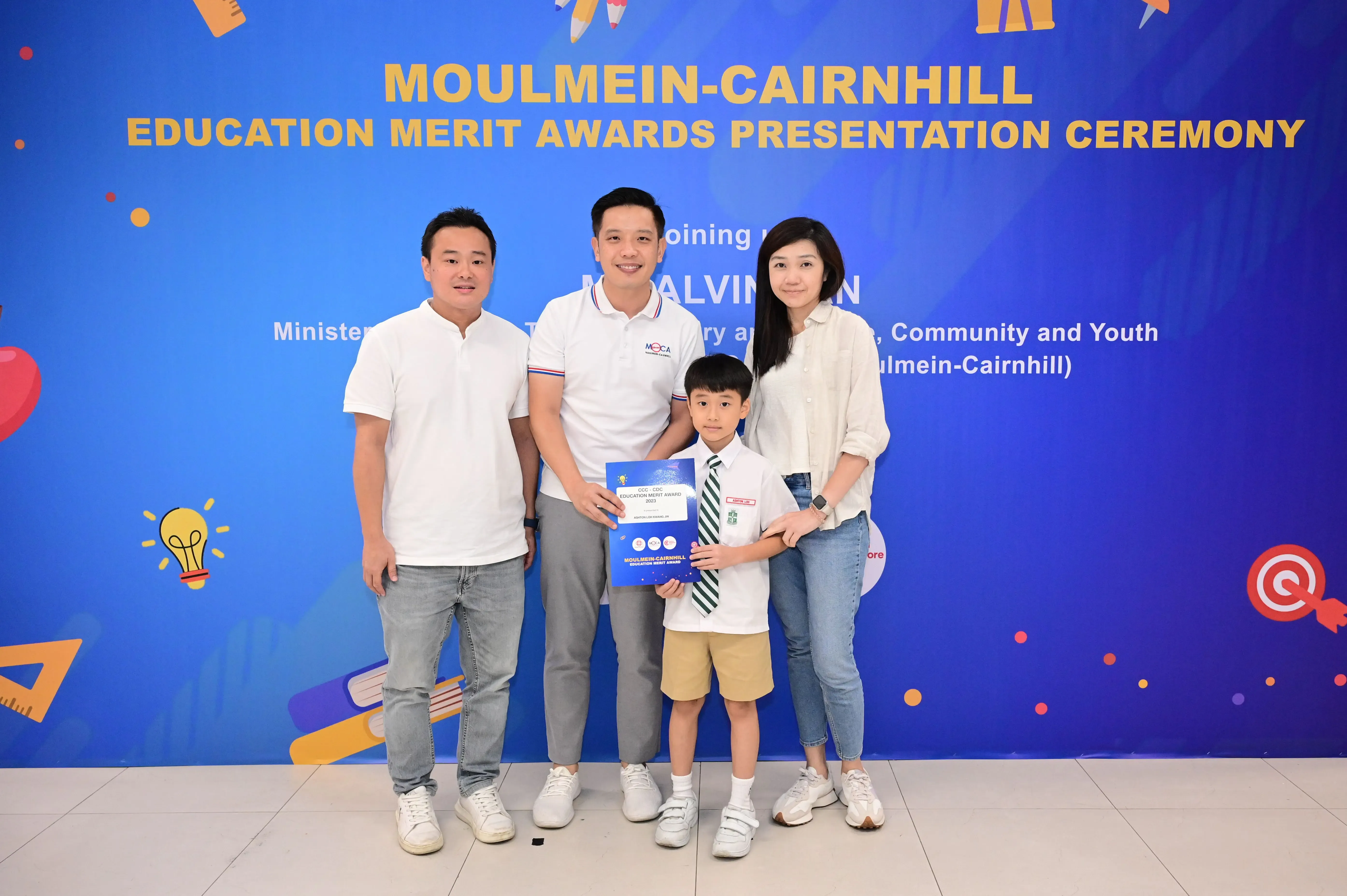 Three adults and a young boy holding an award certificate pose before a Moulmein-Cairnhill Education Merit Awards banner.