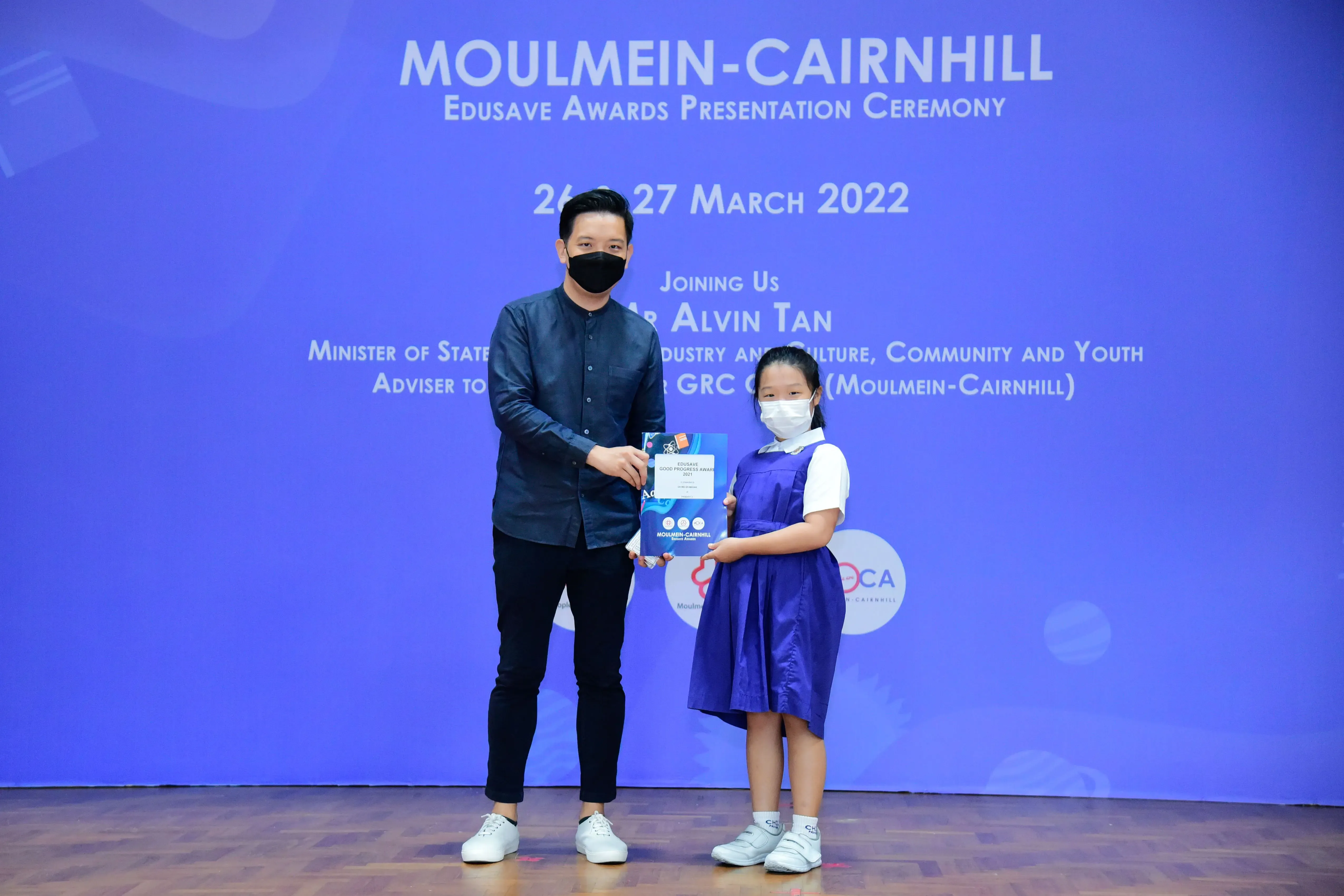 A masked man and a young girl in school uniform hold an award at the Moulmein-Cairnhill Edusave Ceremony, March 2022.