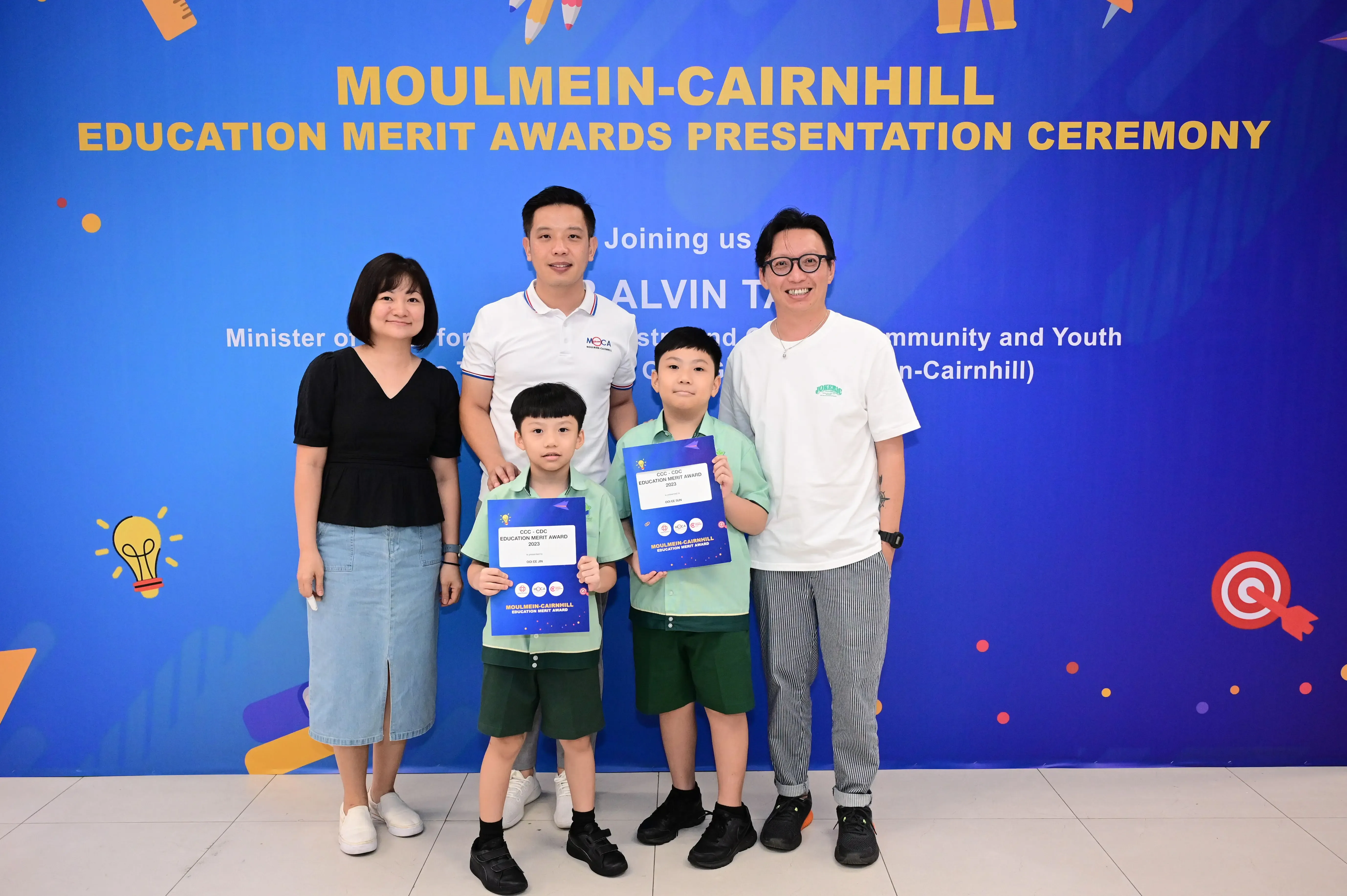 Two boys holding award certificates pose with three adults at the Moulmein-Cairnhill Education Merit Awards ceremony.