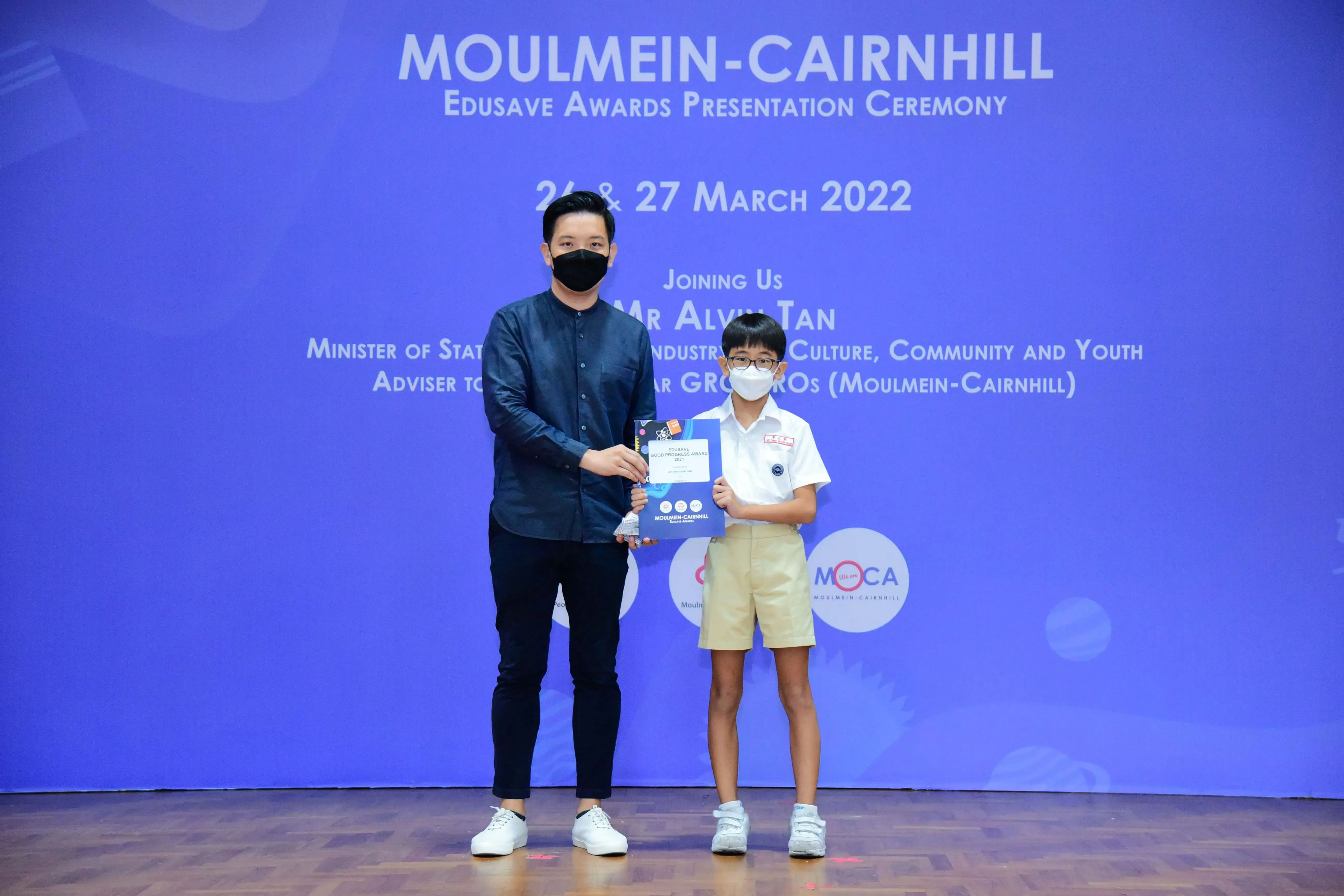 An adult and a young student in school uniform hold a certificate at the Moulmein-Cairnhill Edusave Awards Ceremony 2022.