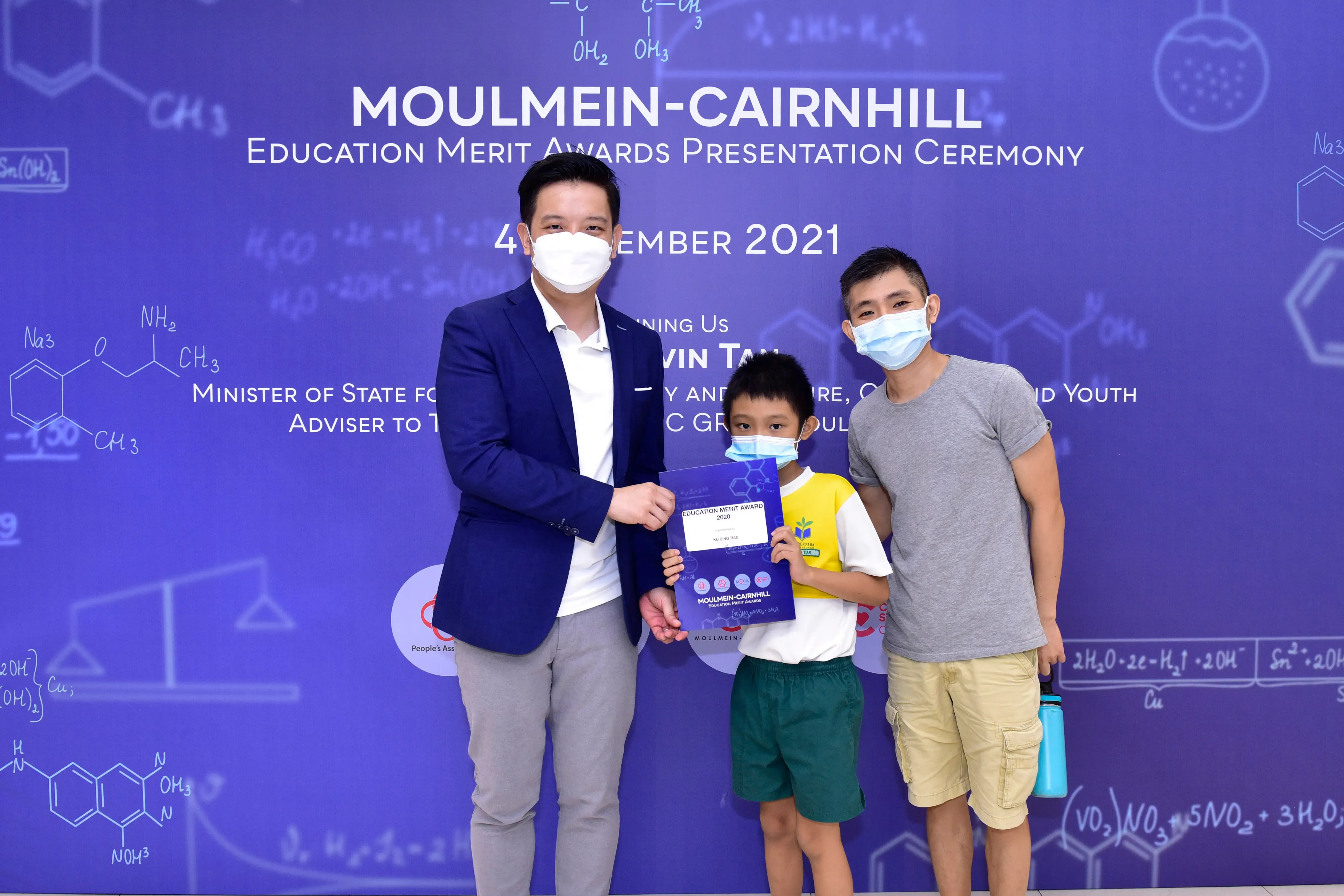 Three masked people pose at the Moulmein-Cairnhill Education Merit Awards 2021, with a boy holding a certificate.
