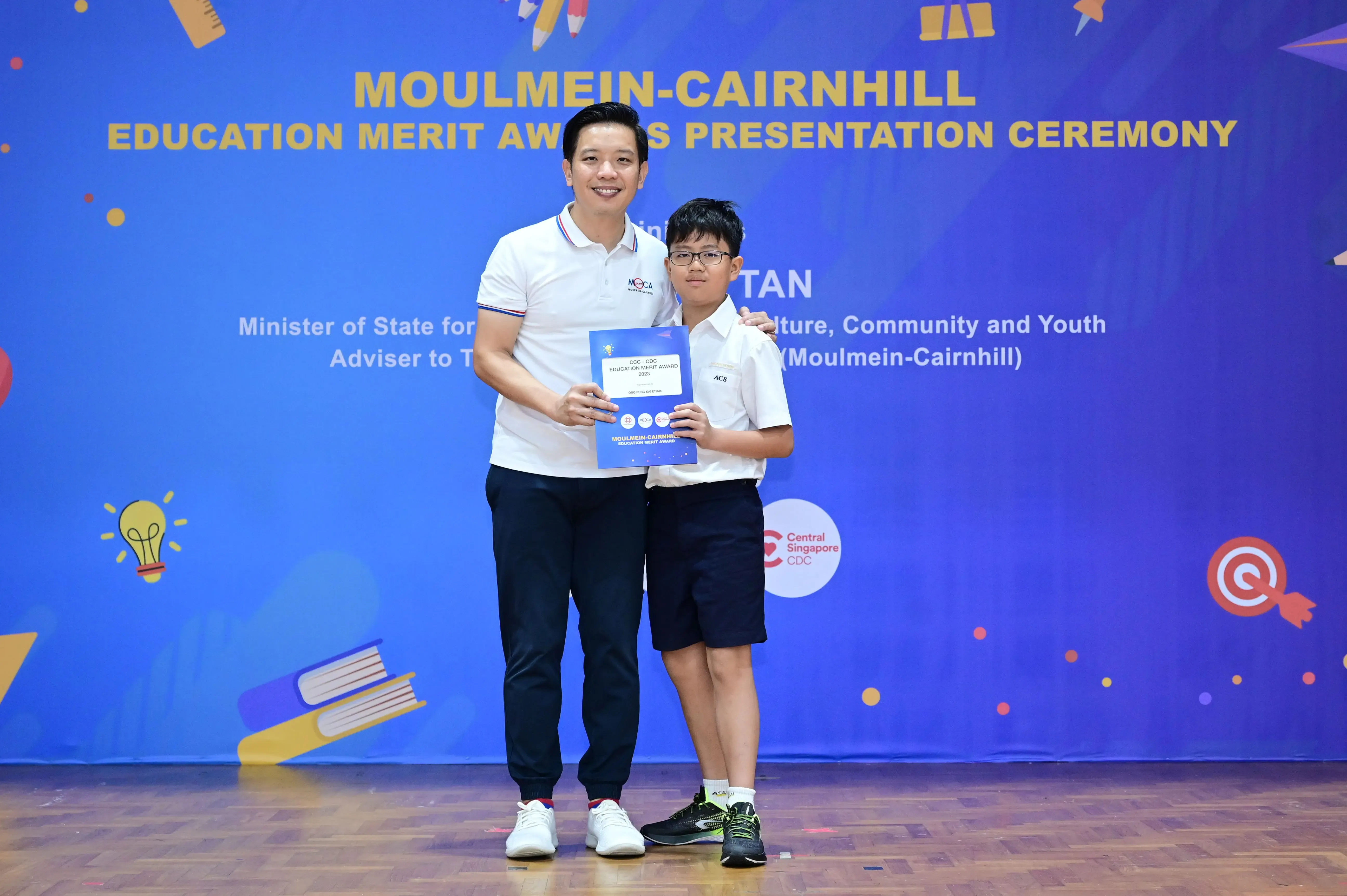 An adult and a young boy holding a certificate at the Moulmein-Cairnhill Education Merit Awards Presentation Ceremony.