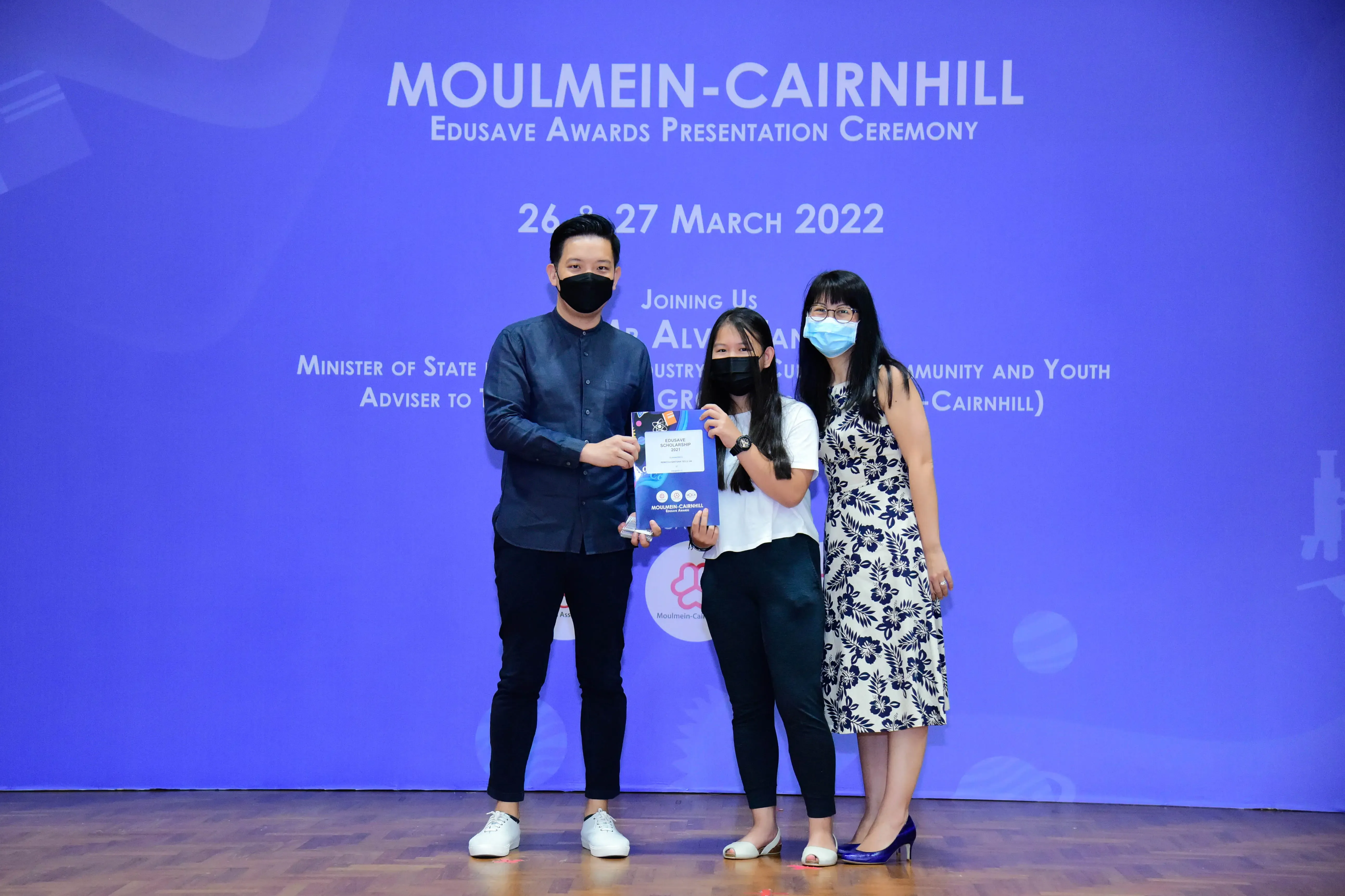 Three masked people stand on a stage holding an award at the Moulmein-Cairnhill Edusave Awards Ceremony, March 2022.
