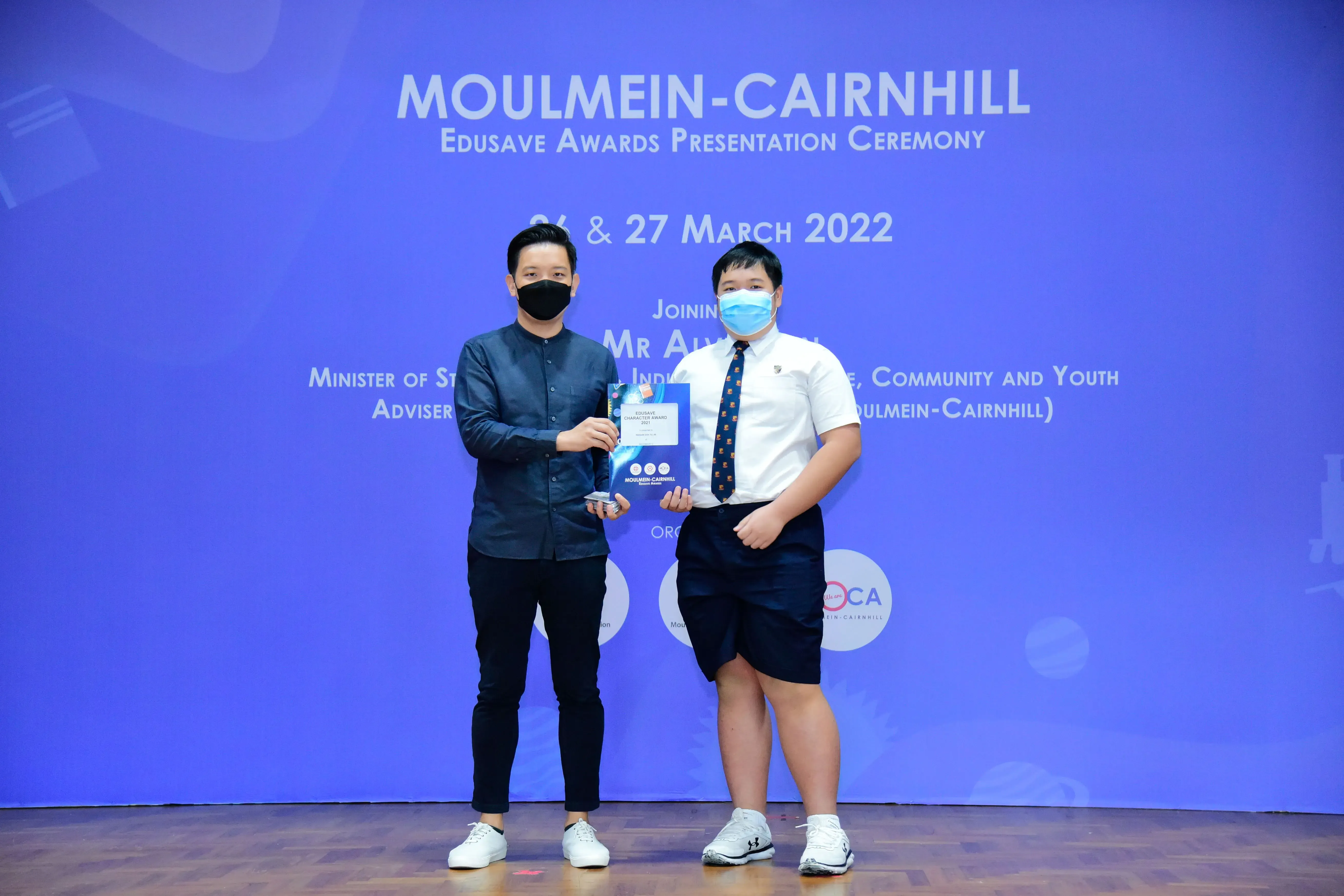 Two masked individuals hold an award at the Moulmein-Cairnhill Edusave Awards Presentation Ceremony, March 2022.