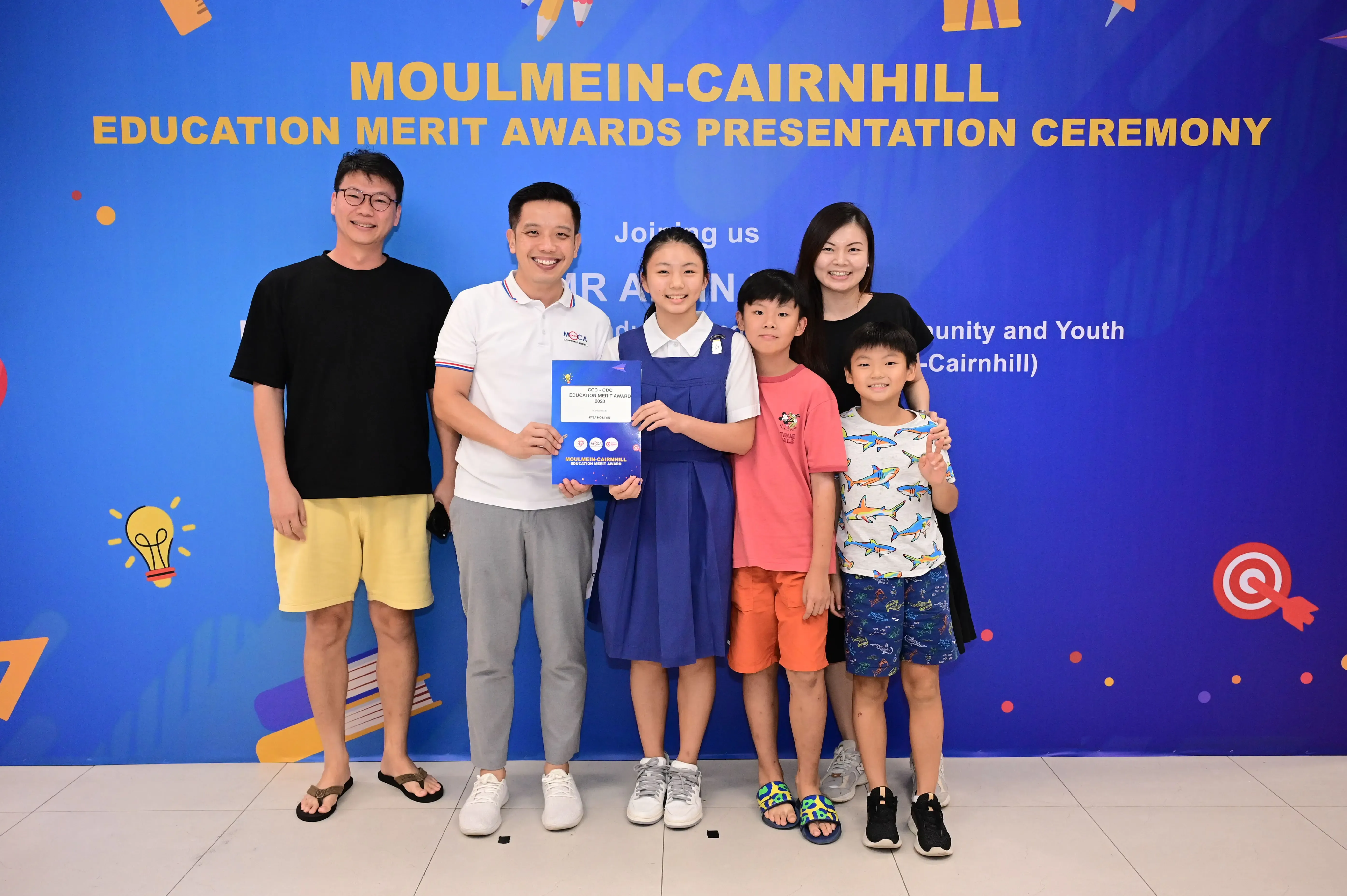 Six people pose before a Moulmein-Cairnhill Education Merit Awards banner, with a girl holding a blue award certificate.