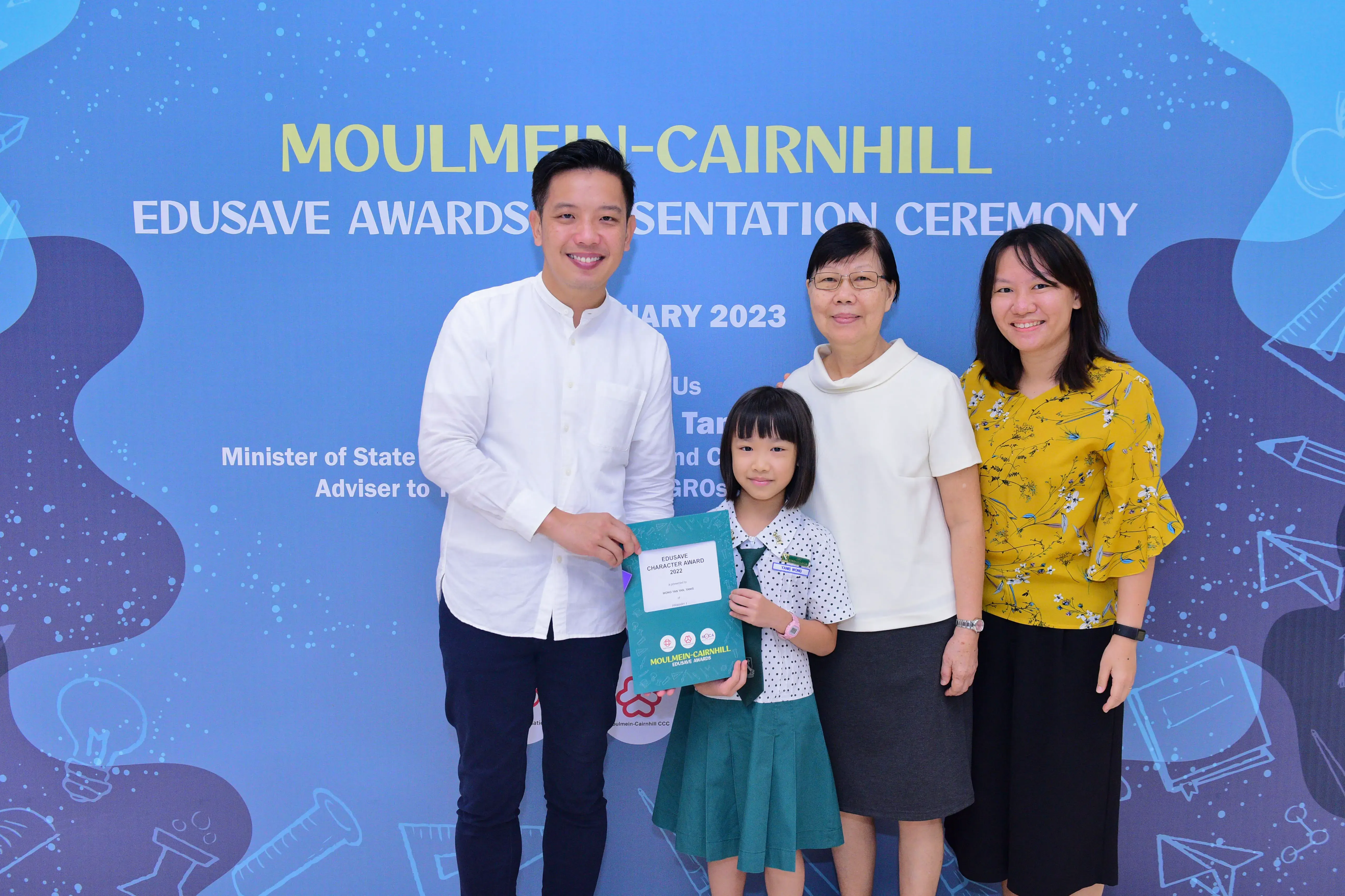 Four people pose at the 2023 Moulmein-Cairnhill Edusave Awards ceremony, with a girl holding a teal award folder.