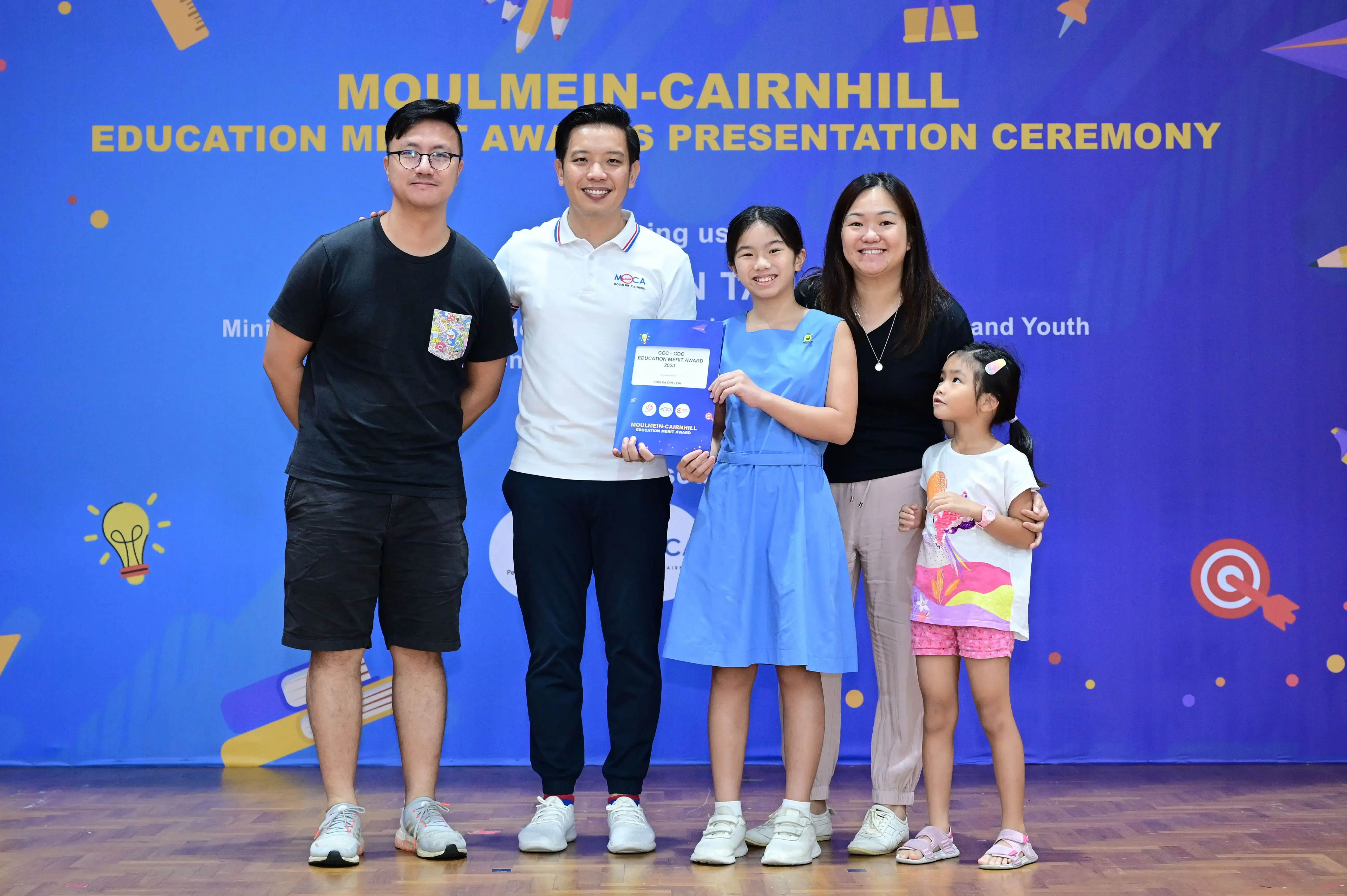 Five people pose at the Moulmein-Cairnhill Education Merit Awards Presentation Ceremony, with a girl holding an award certificate.