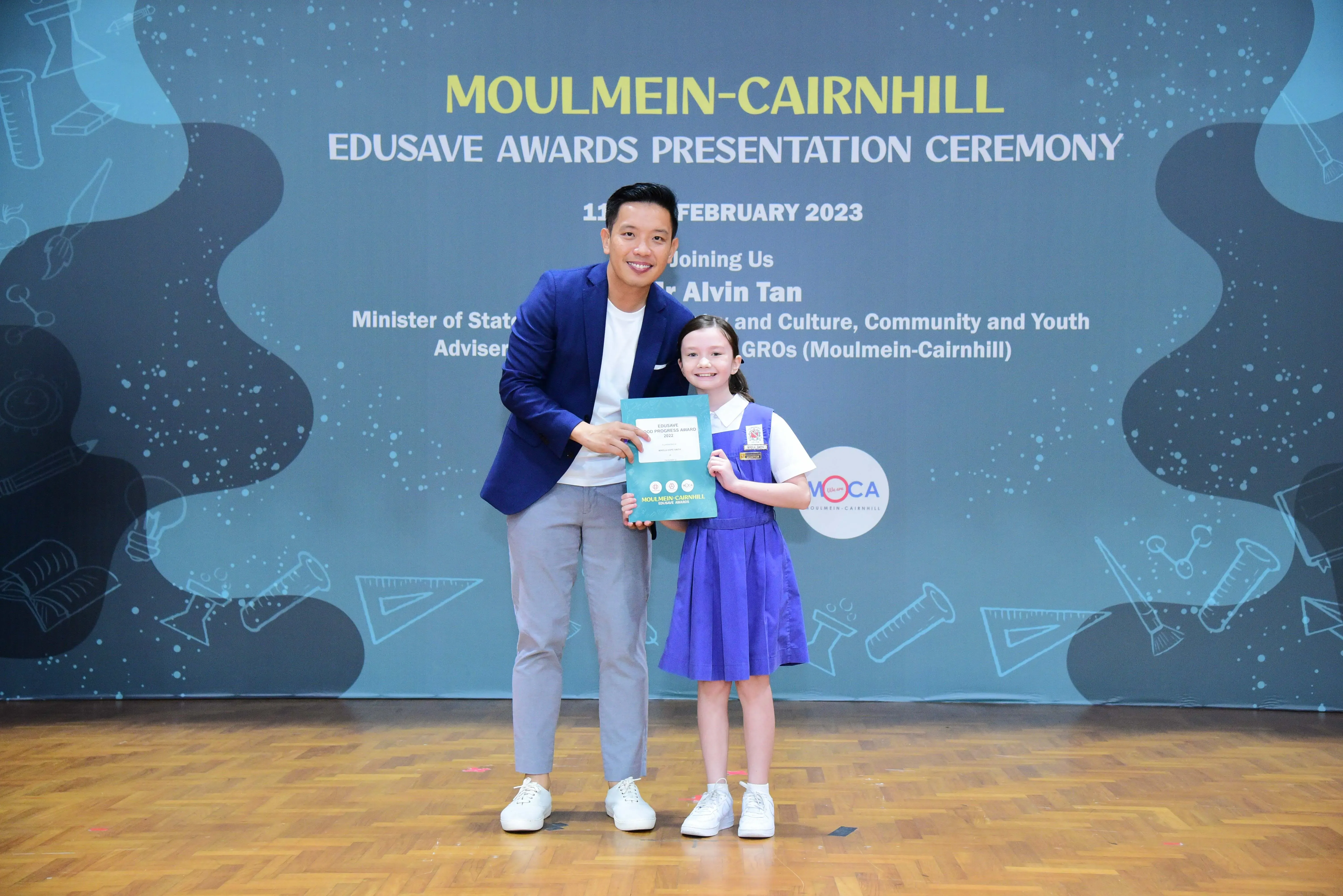 A man in a blue blazer and a young girl in a school uniform hold a certificate at the Moulmein-Cairnhill Edusave Awards Ceremony.