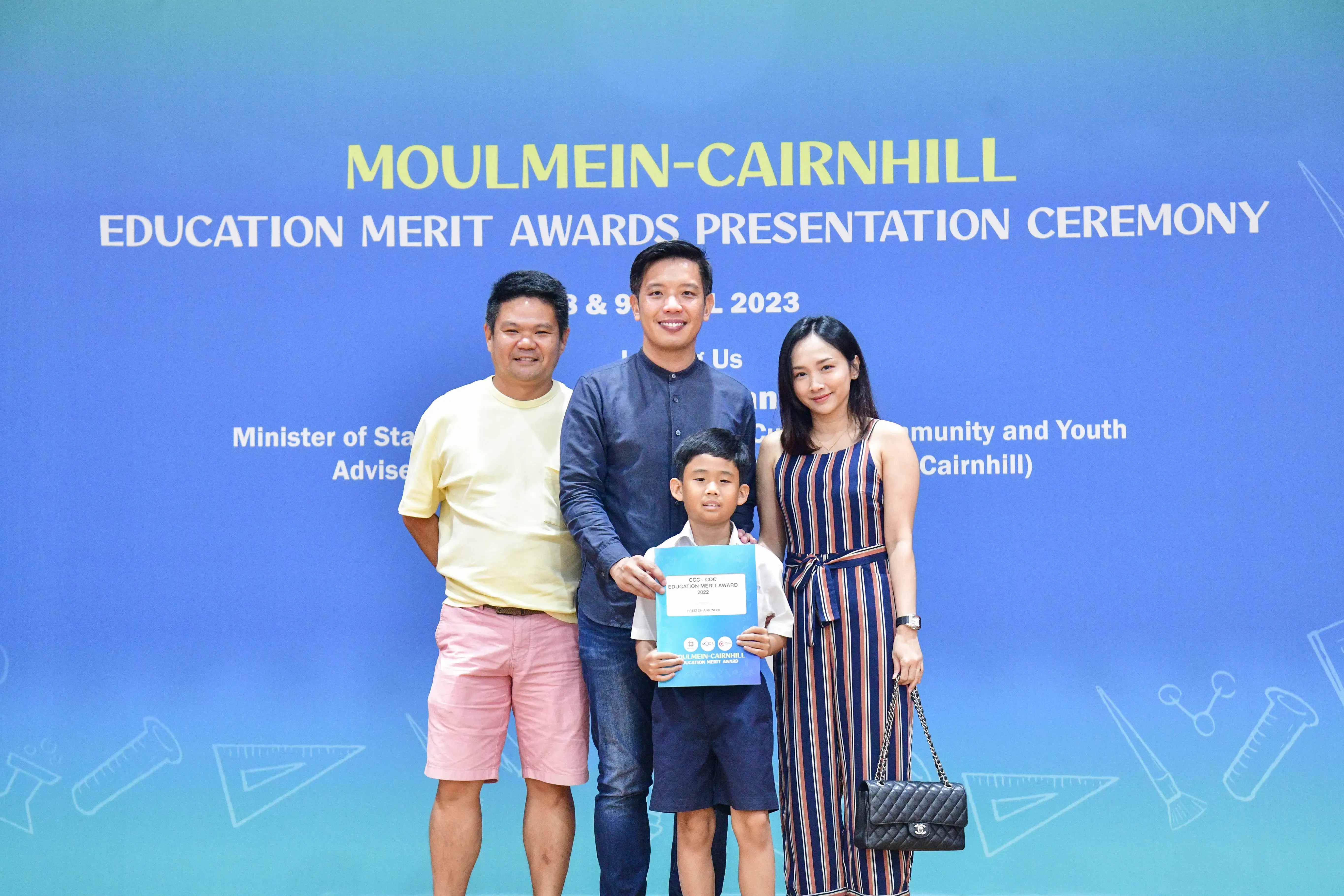 Three adults and a boy holding an award certificate pose before a Moulmein-Cairnhill Education Merit Awards banner.