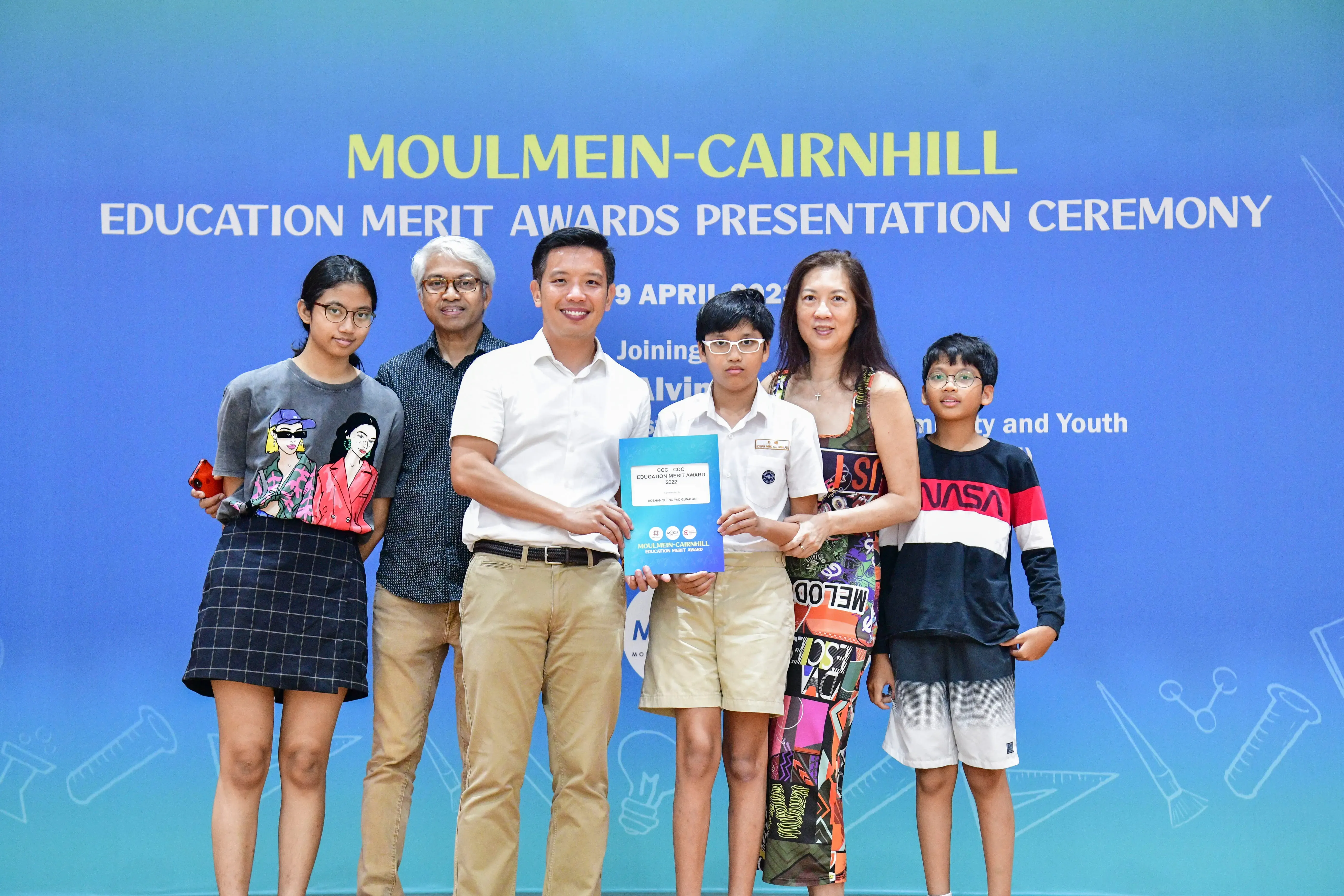 Six people pose at the Moulmein-Cairnhill Education Merit Awards Presentation Ceremony, with a boy holding a blue award folder.