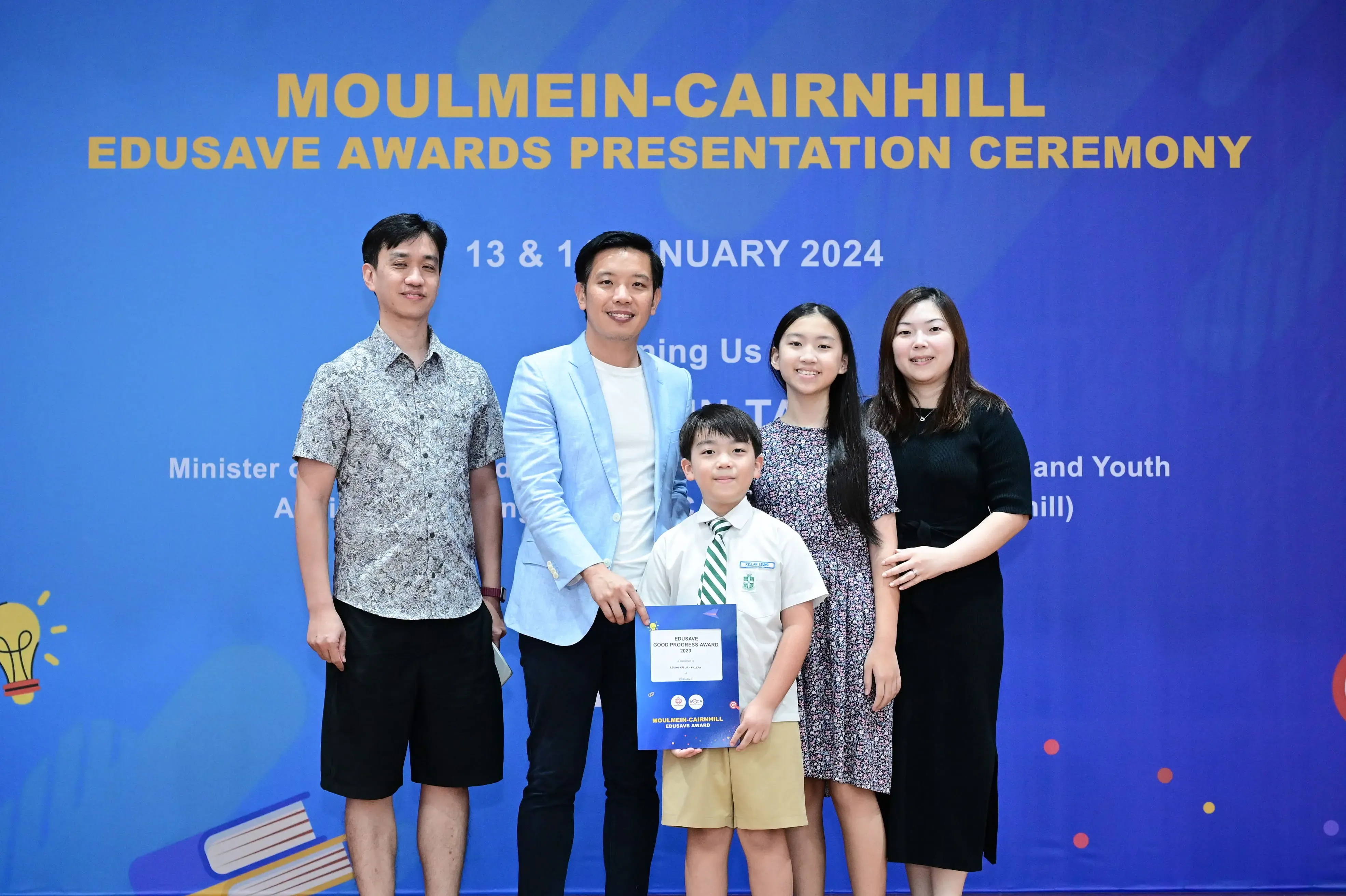 Five people pose before a blue banner reading Moulmein-Cairnhill Edusave Awards Presentation Ceremony 2024; a boy holds a certificate.