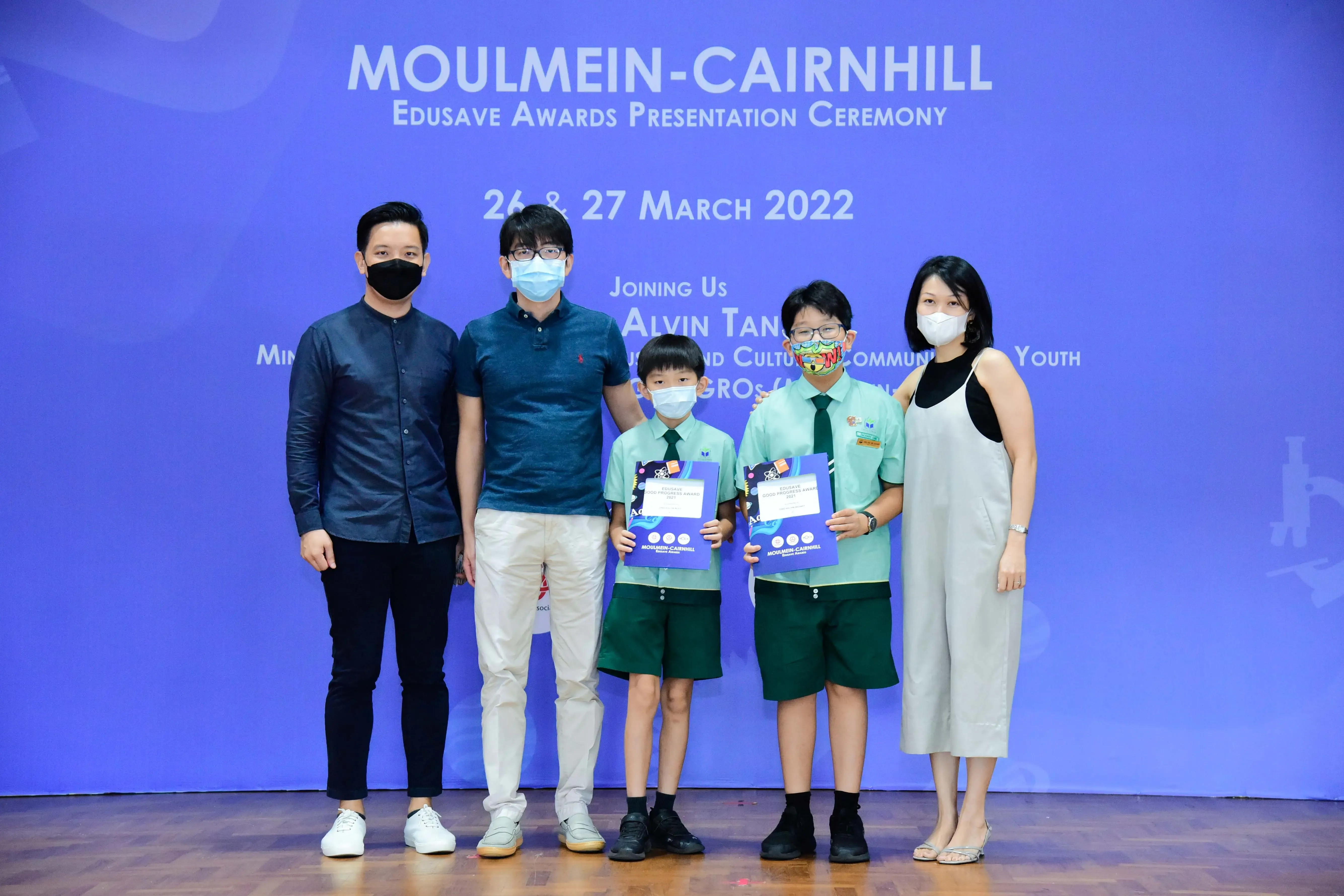 Five masked people pose at the Moulmein-Cairnhill Edusave Awards Ceremony on 26-27 March 2022, two children holding award folders.