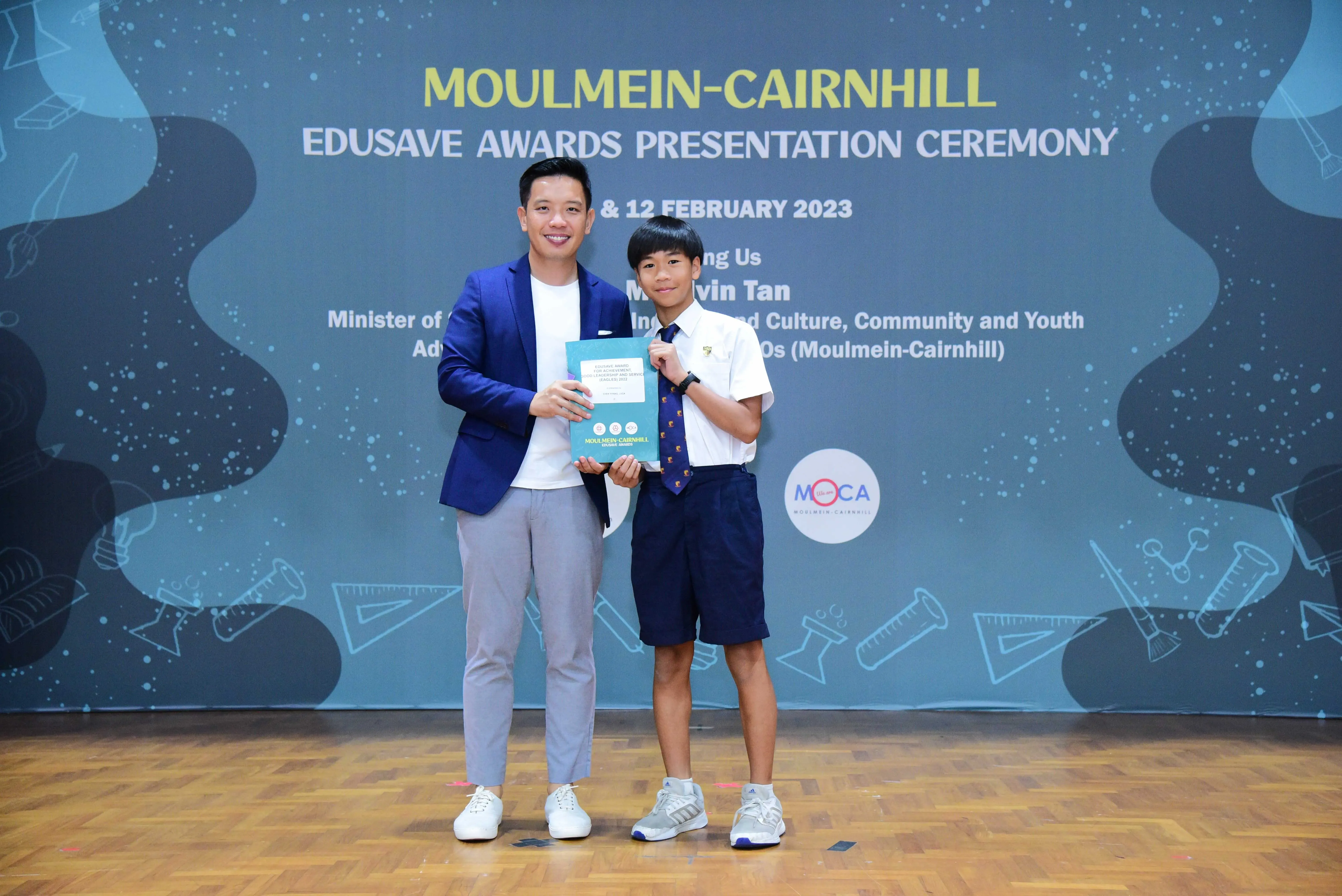 Man in blue blazer and young boy in school uniform hold a booklet at Moulmein-Cairnhill Edusave Awards Ceremony 2023.