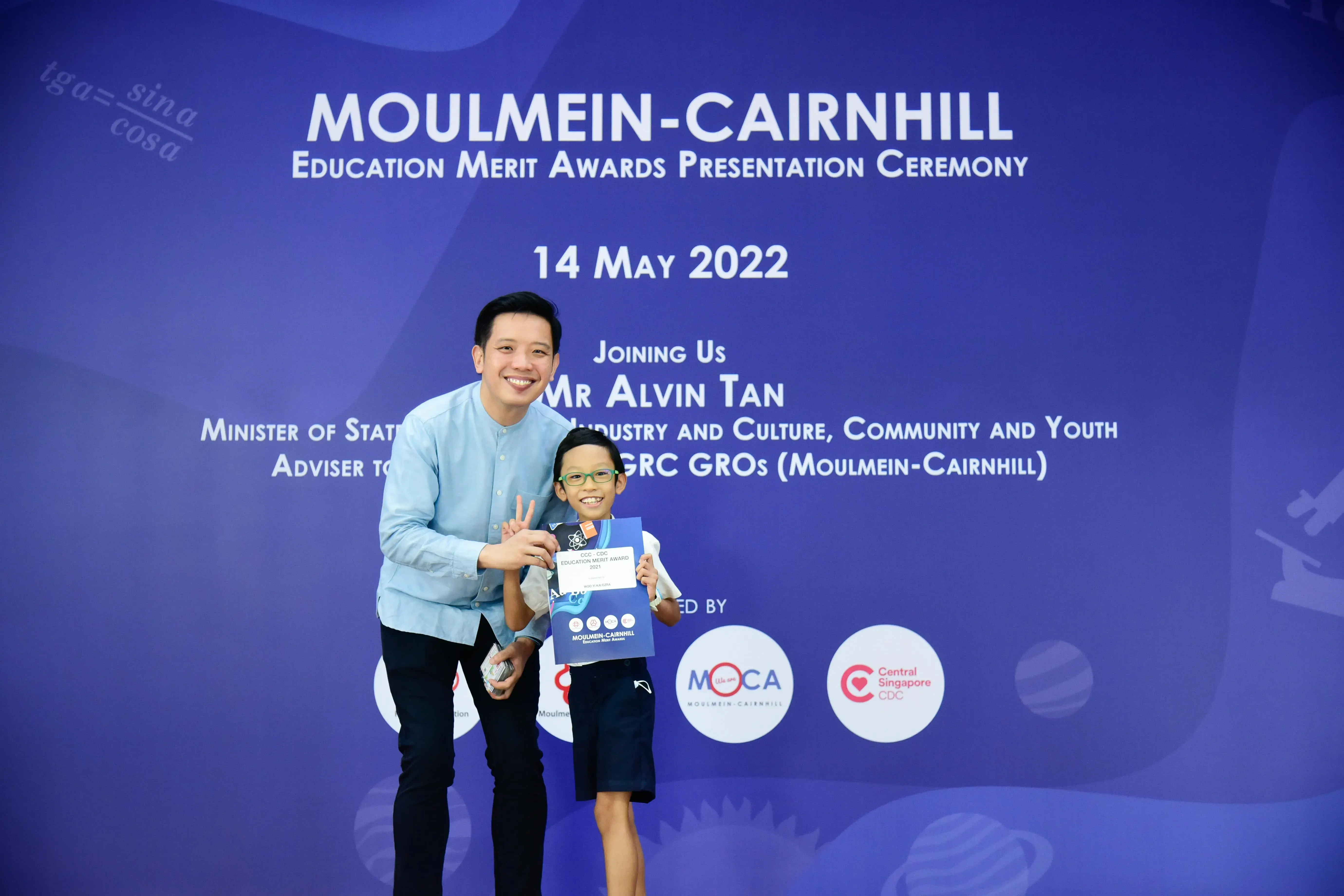 A man and a young boy holding an award certificate pose in front of a Moulmein-Cairnhill Education Merit Awards banner.