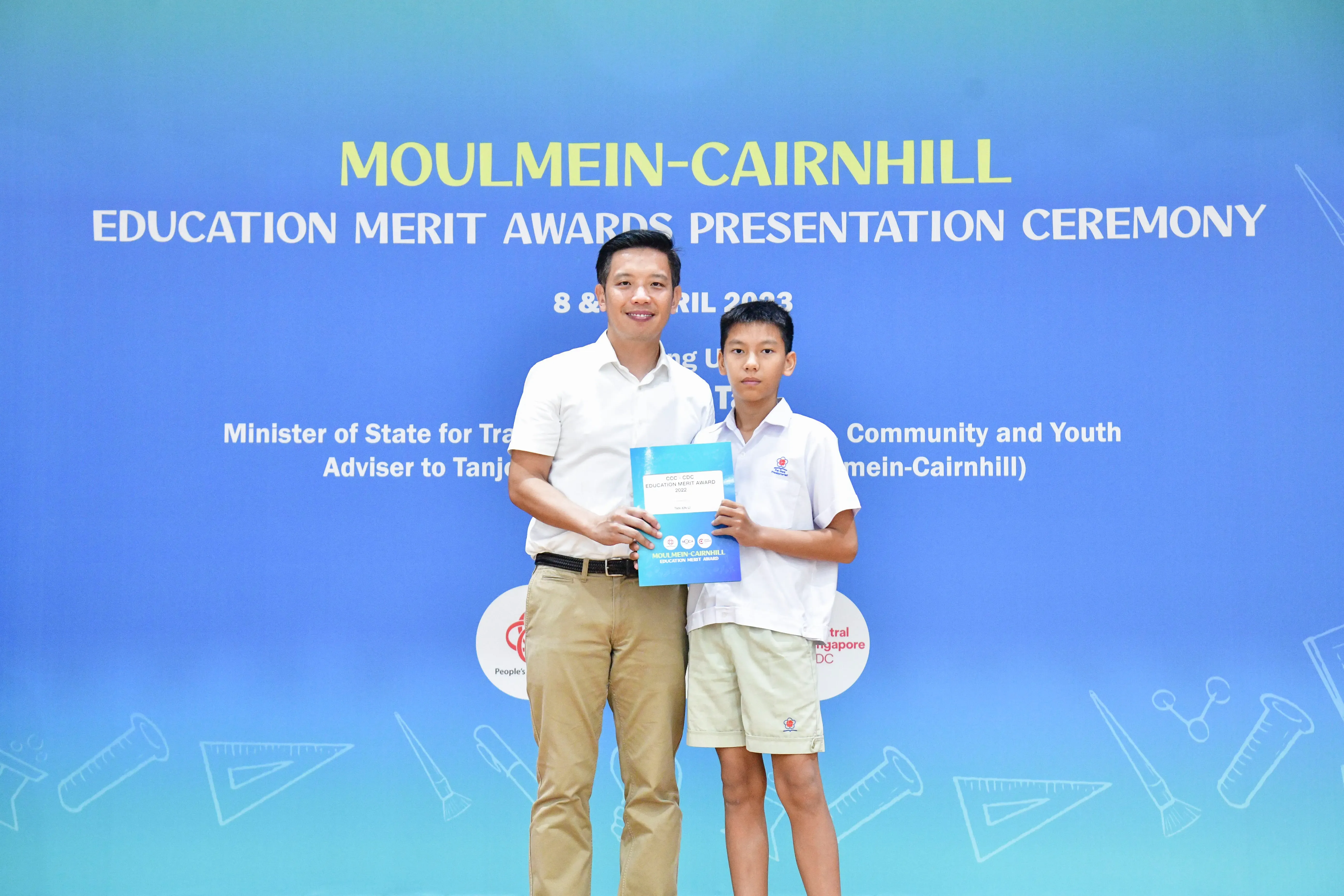 An adult and a young student hold a certificate together at the Moulmein-Cairnhill Education Merit Awards Ceremony.