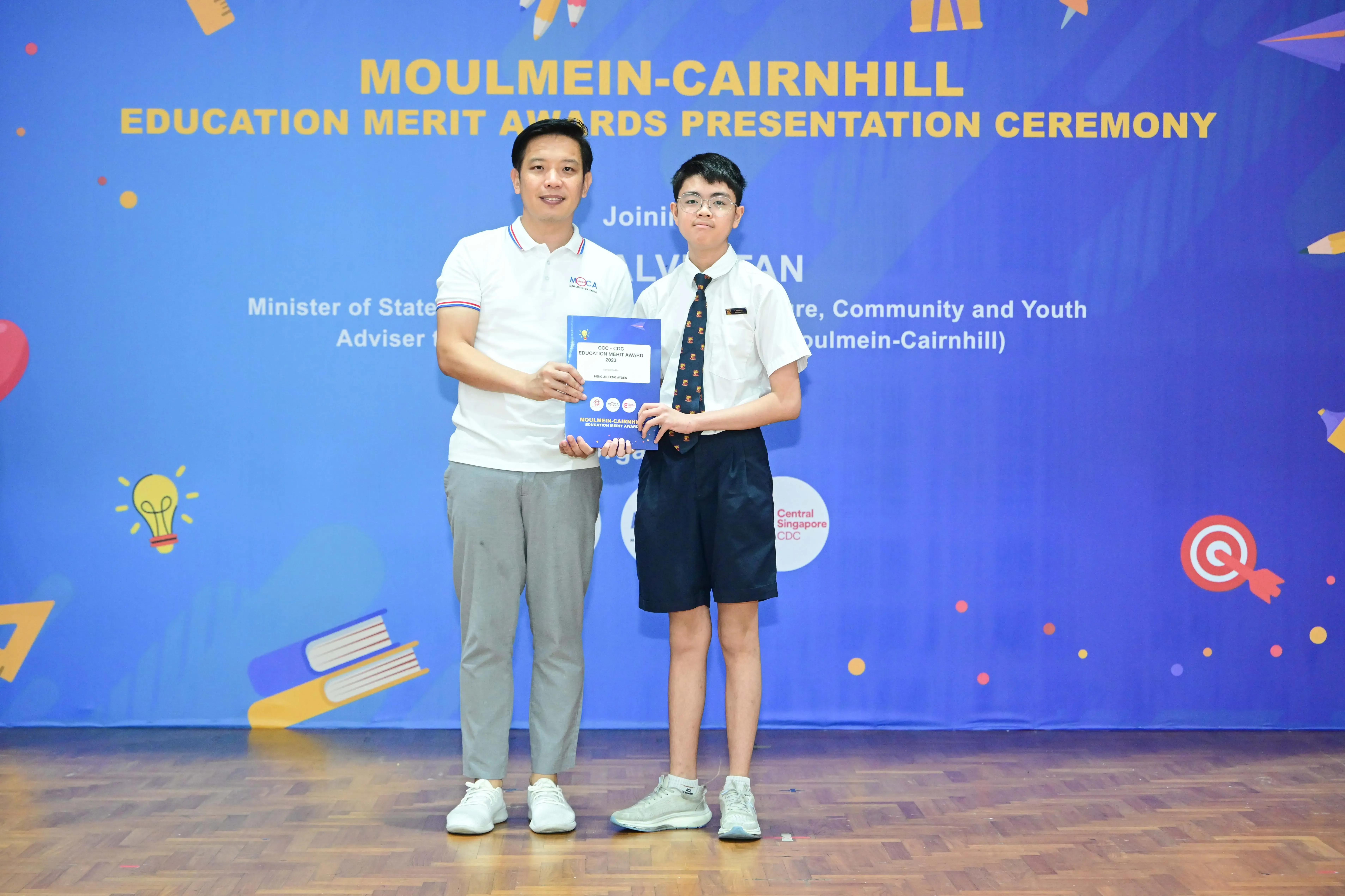 Two people hold a certificate together at the Moulmein-Cairnhill Education Merit Awards Presentation Ceremony.