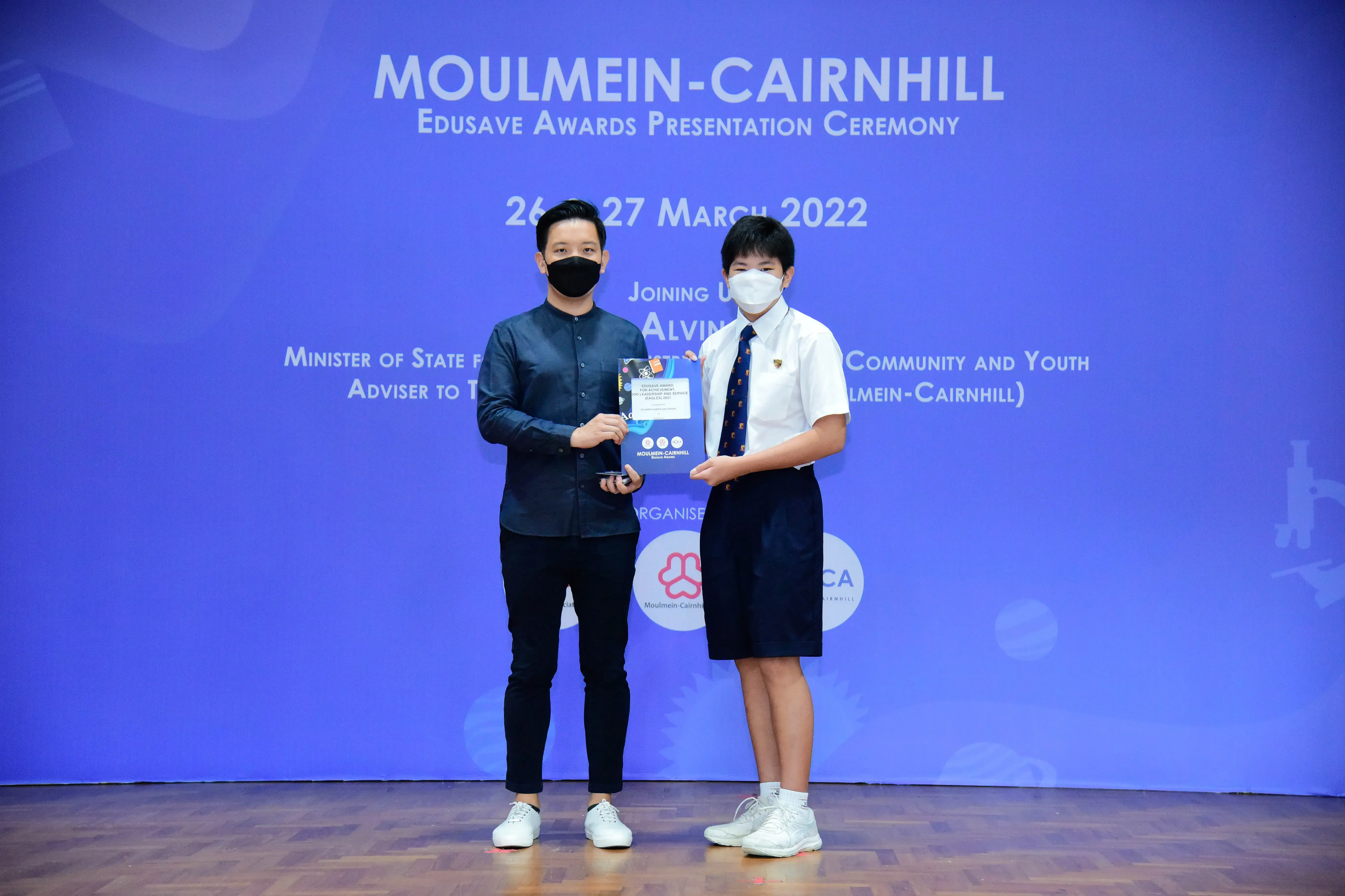 Two masked individuals hold an award at the Moulmein-Cairnhill Edusave Awards Presentation Ceremony, March 2022.