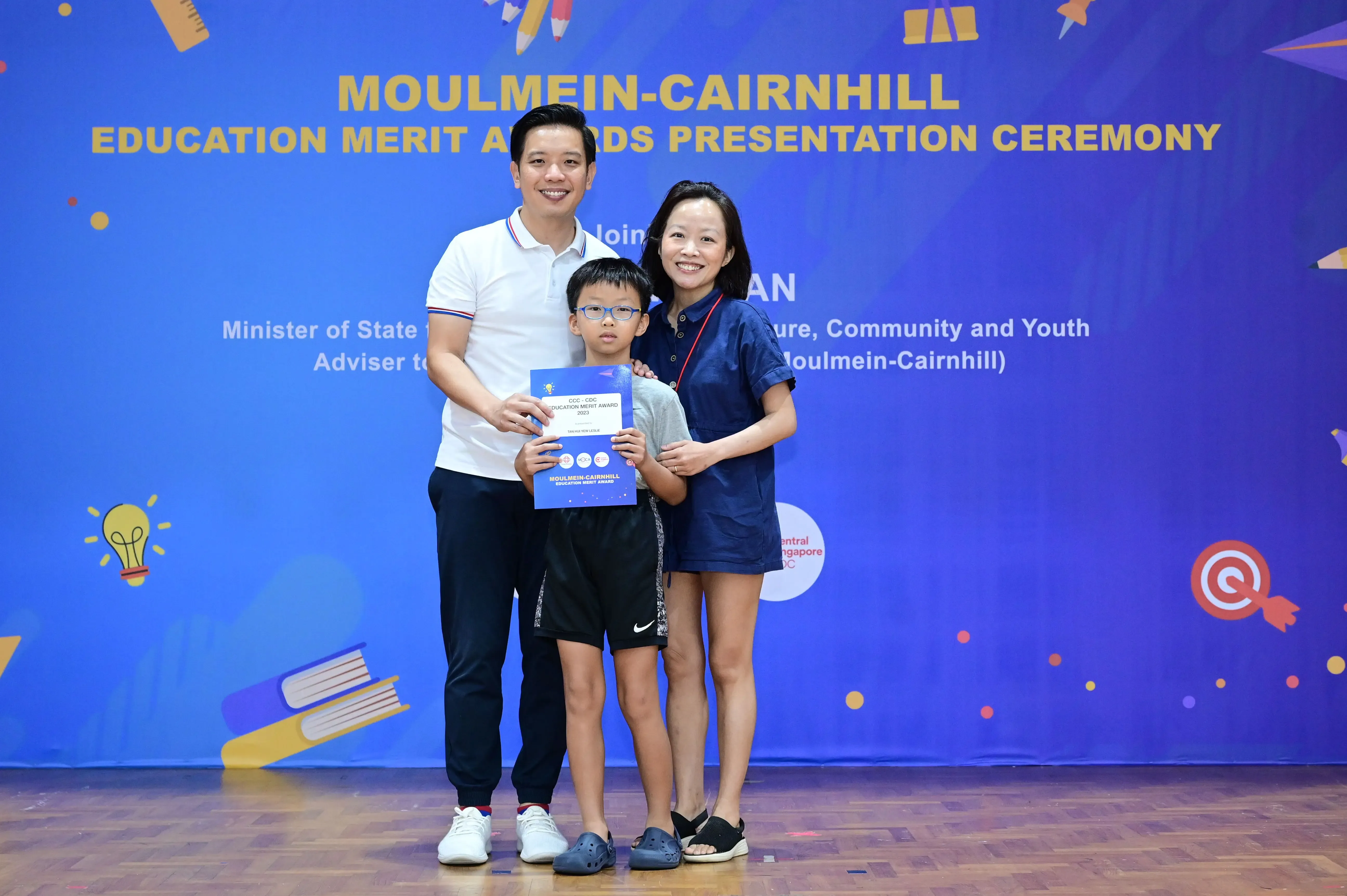A boy holding an award certificate stands with two smiling adults at the Moulmein-Cairnhill Education Merit Awards ceremony.