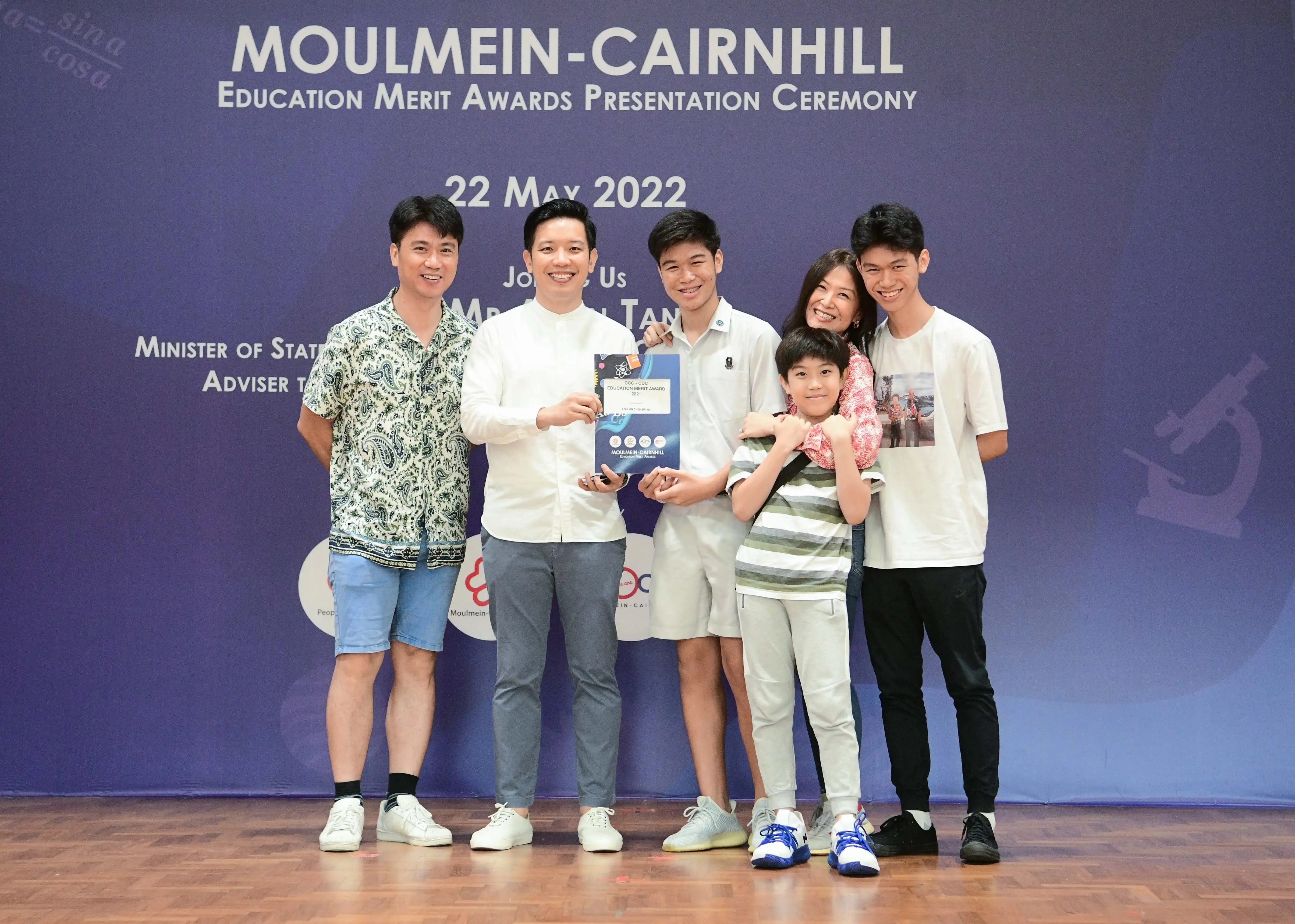 Six people pose smiling in front of a Moulmein-Cairnhill Education Merit Awards banner dated 22 May 2022.