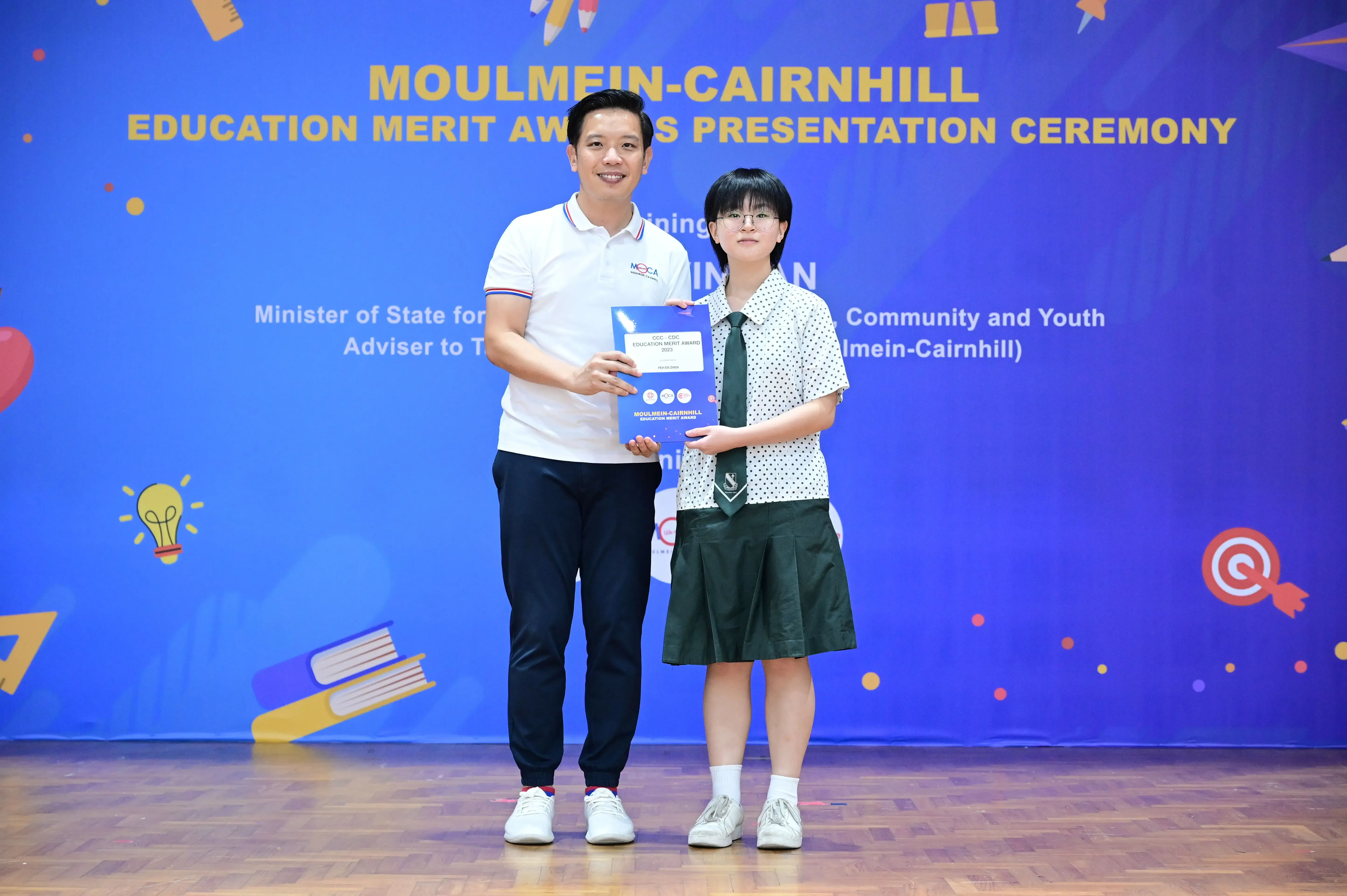 A man and a uniformed student hold an award certificate at the Moulmein-Cairnhill Education Merit Awards ceremony.