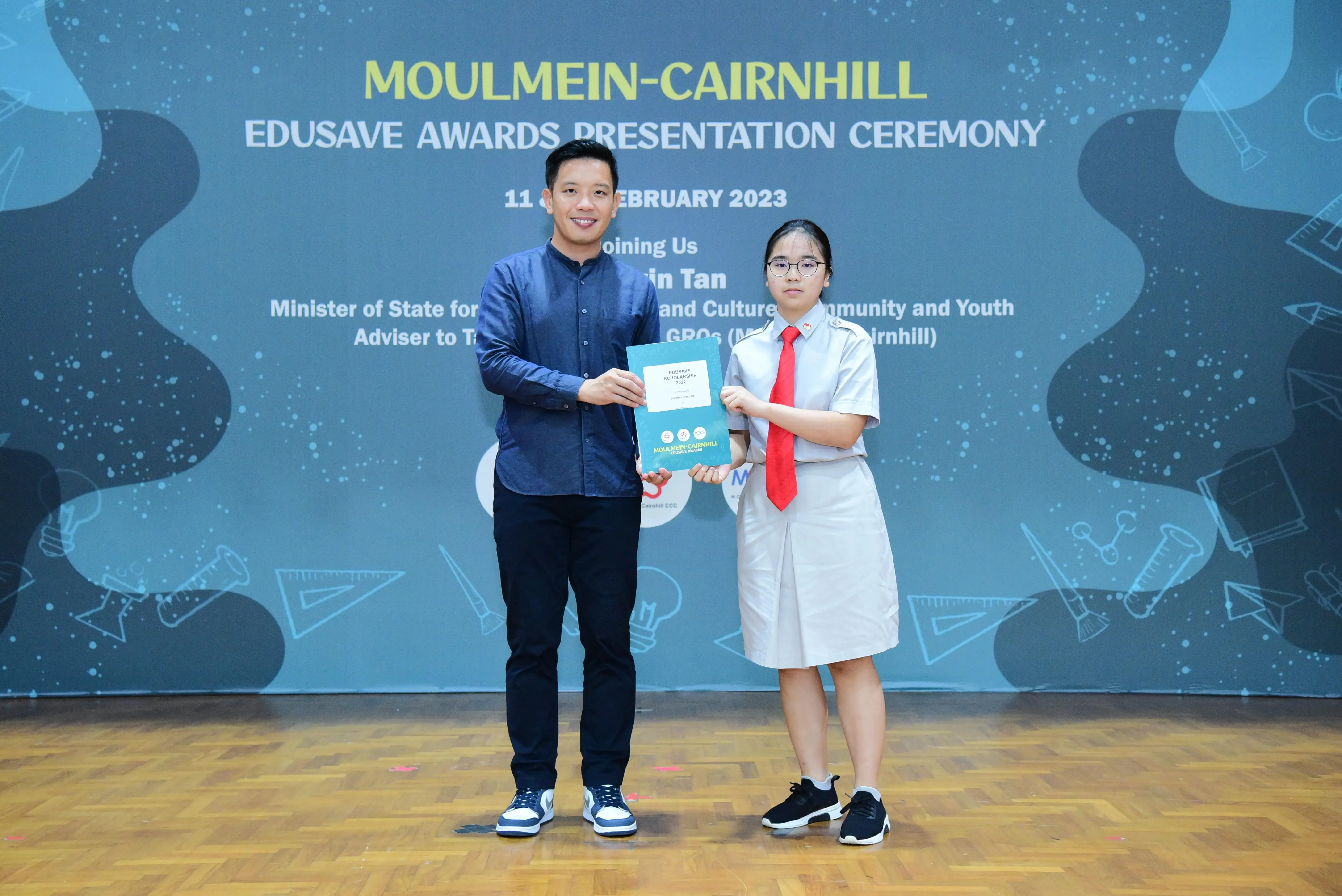 A man and a young girl in school uniform hold a certificate at the Moulmein-Cairnhill Edusave Awards Ceremony 2023.