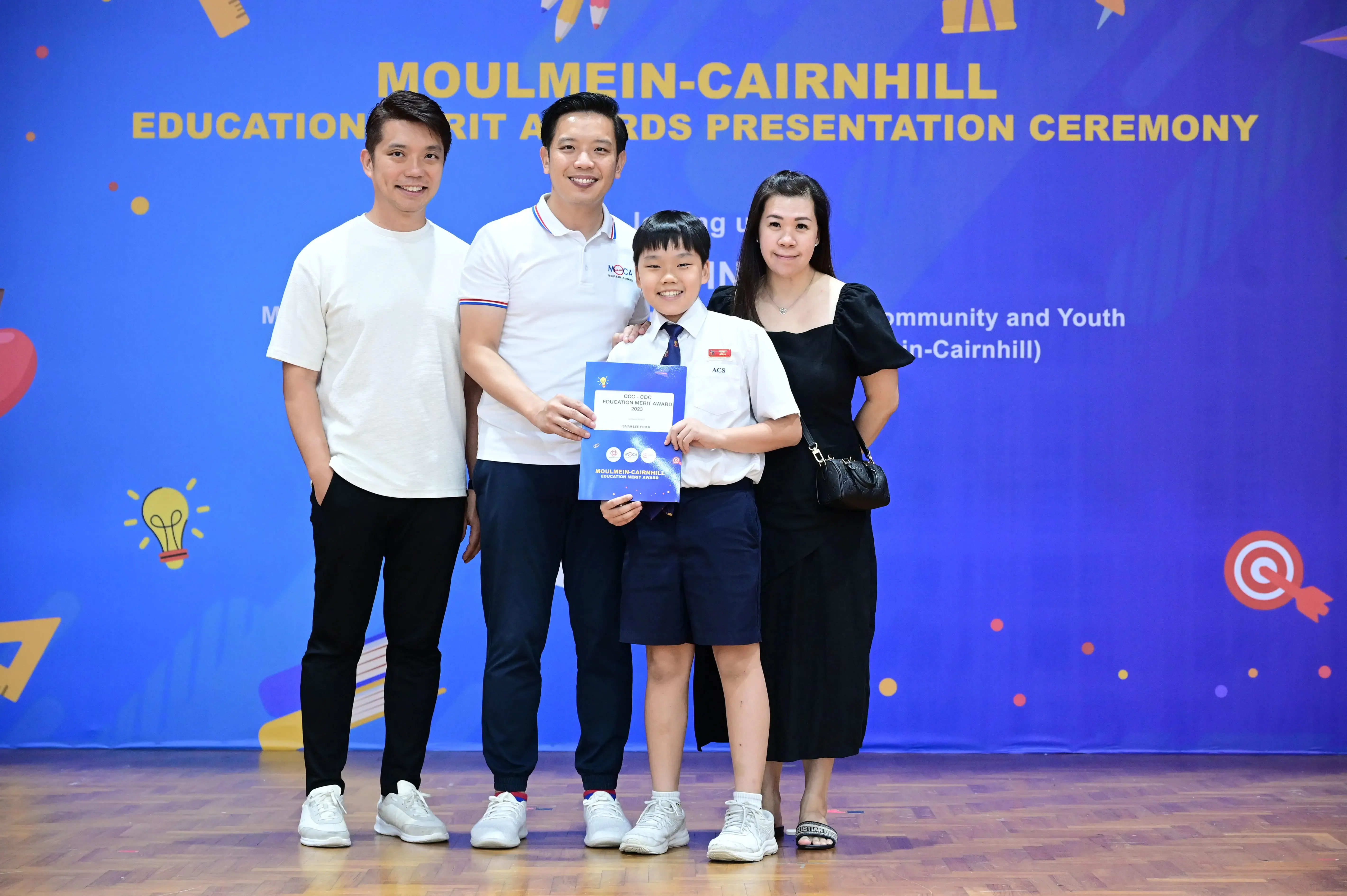 A child in school uniform holds an award certificate with three adults at the Moulmein-Cairnhill Education Merit Awards ceremony.