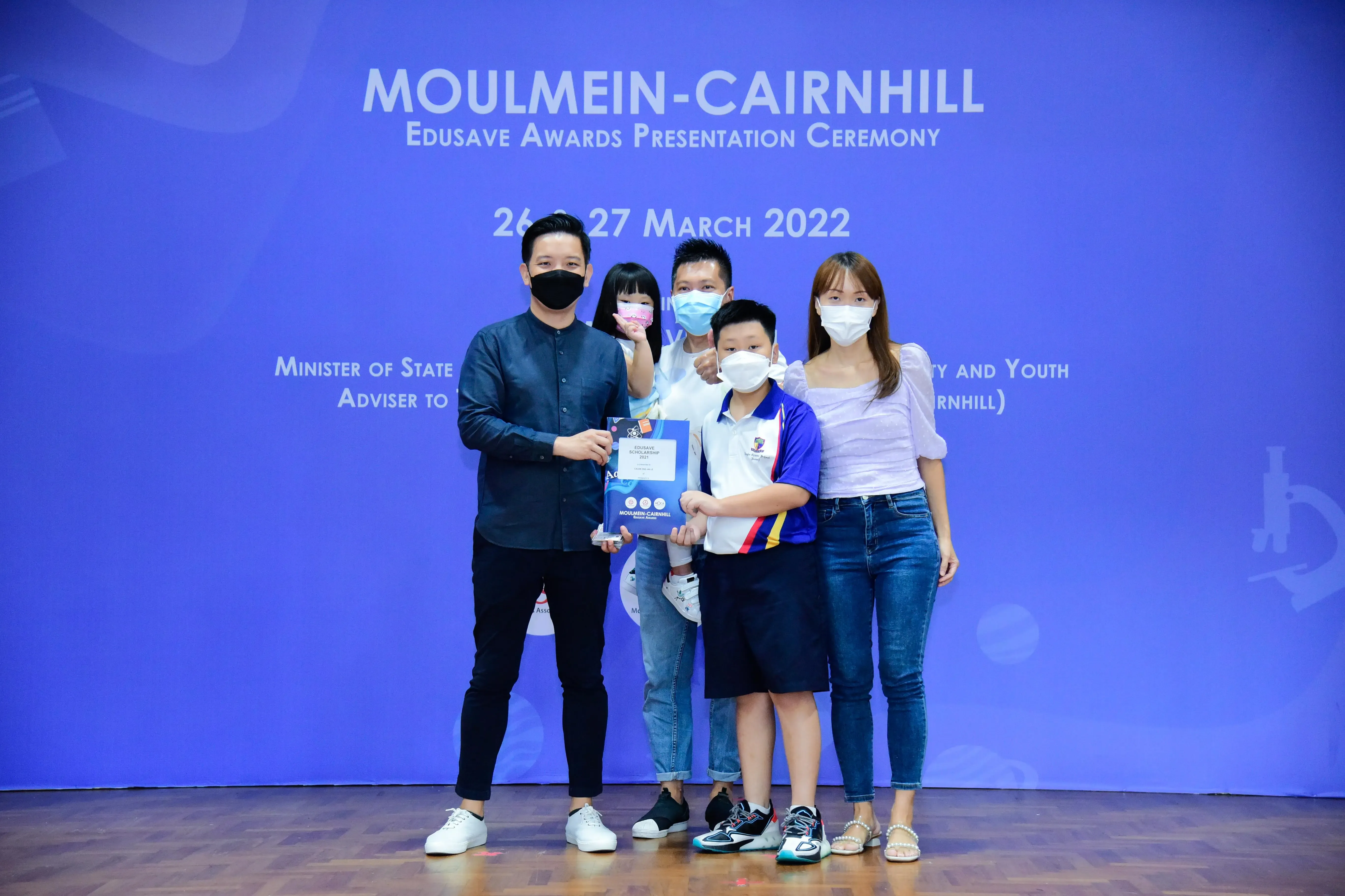 Four masked people pose with a child holding an award at the Moulmein-Cairnhill Edusave Awards Ceremony, March 2022.