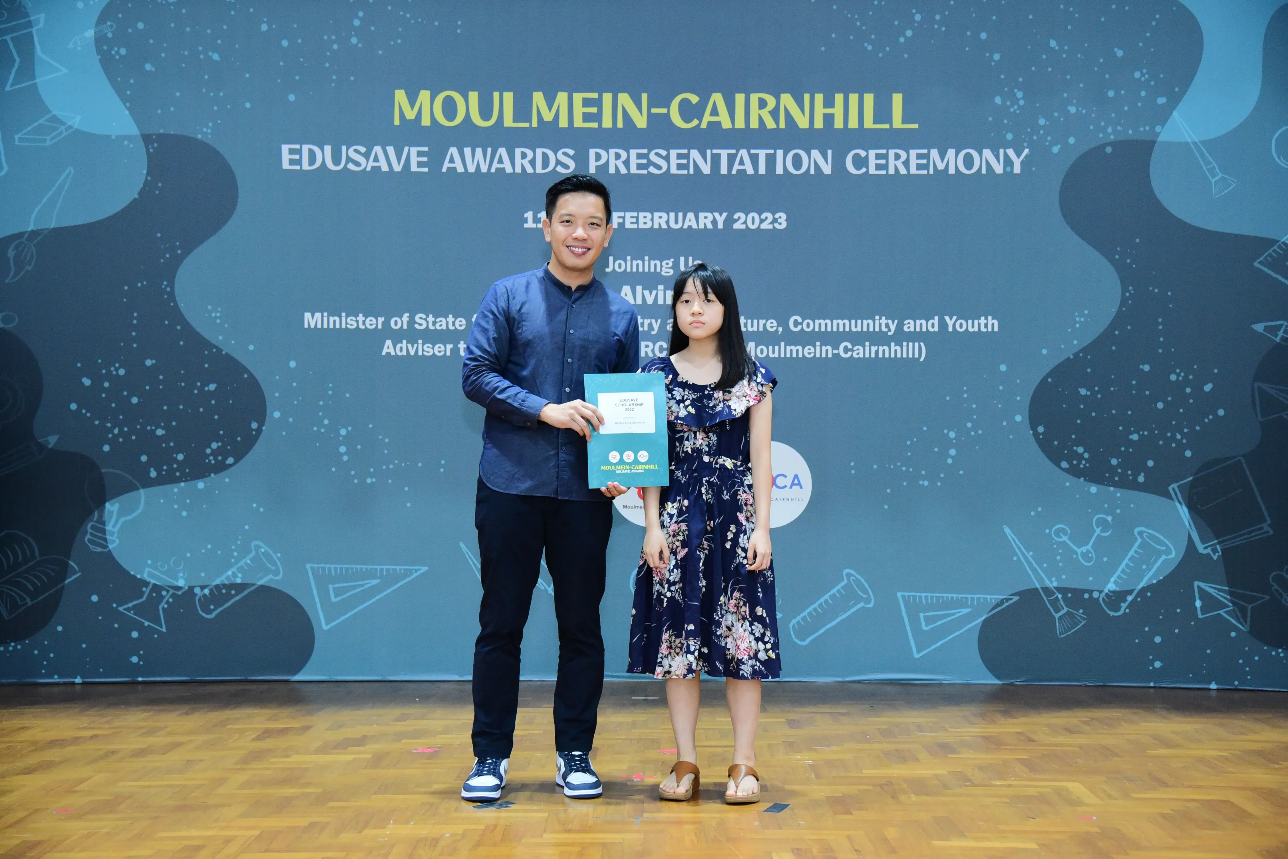 A man and a young girl hold a certificate on stage at the Moulmein-Cairnhill Edusave Awards Ceremony, February 2023.