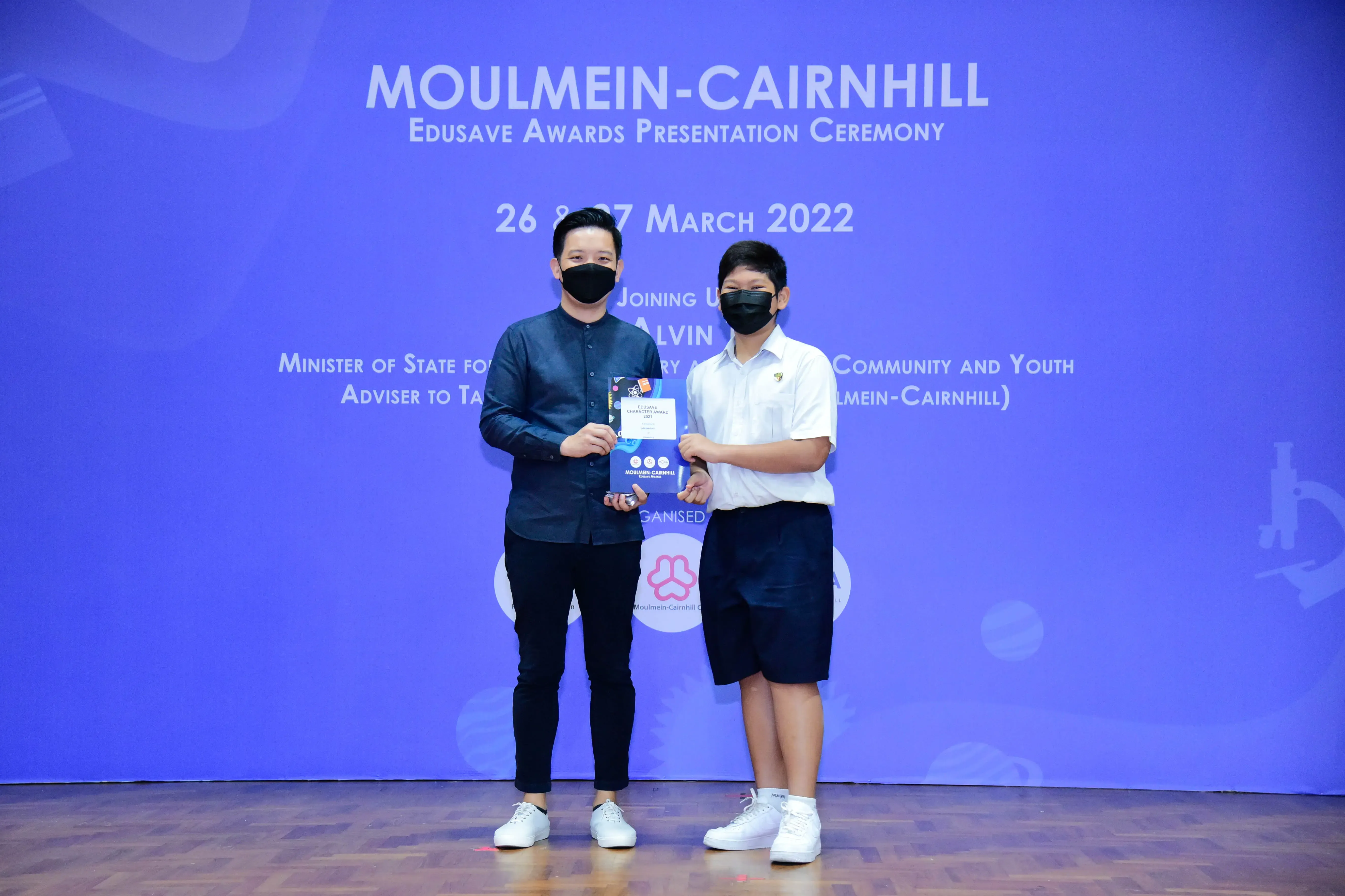Two masked individuals hold an award certificate on stage at the Moulmein-Cairnhill Edusave Awards Ceremony, March 2022.