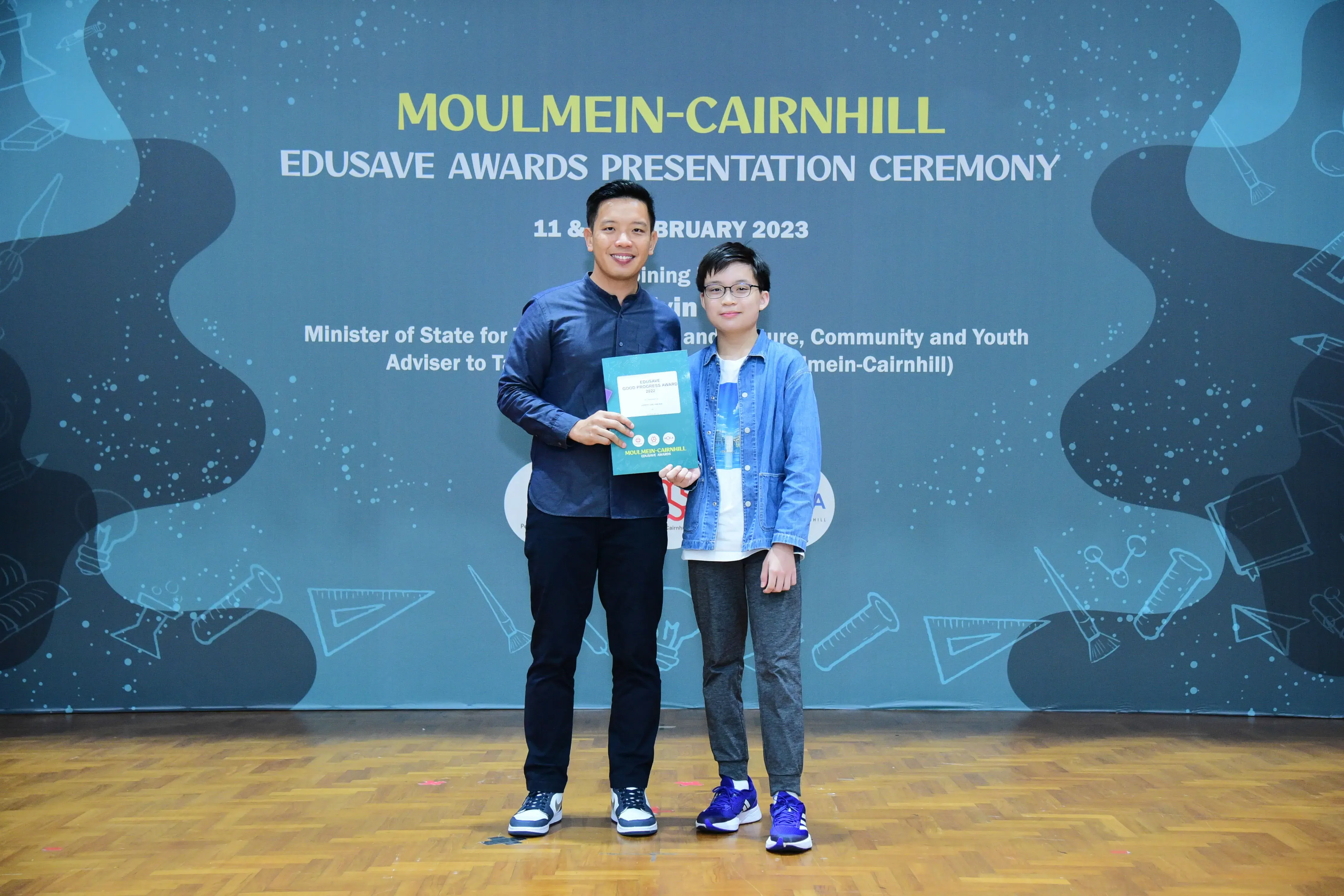 Two people stand before a Moulmein-Cairnhill Edusave Awards Presentation Ceremony backdrop, one holding a certificate.