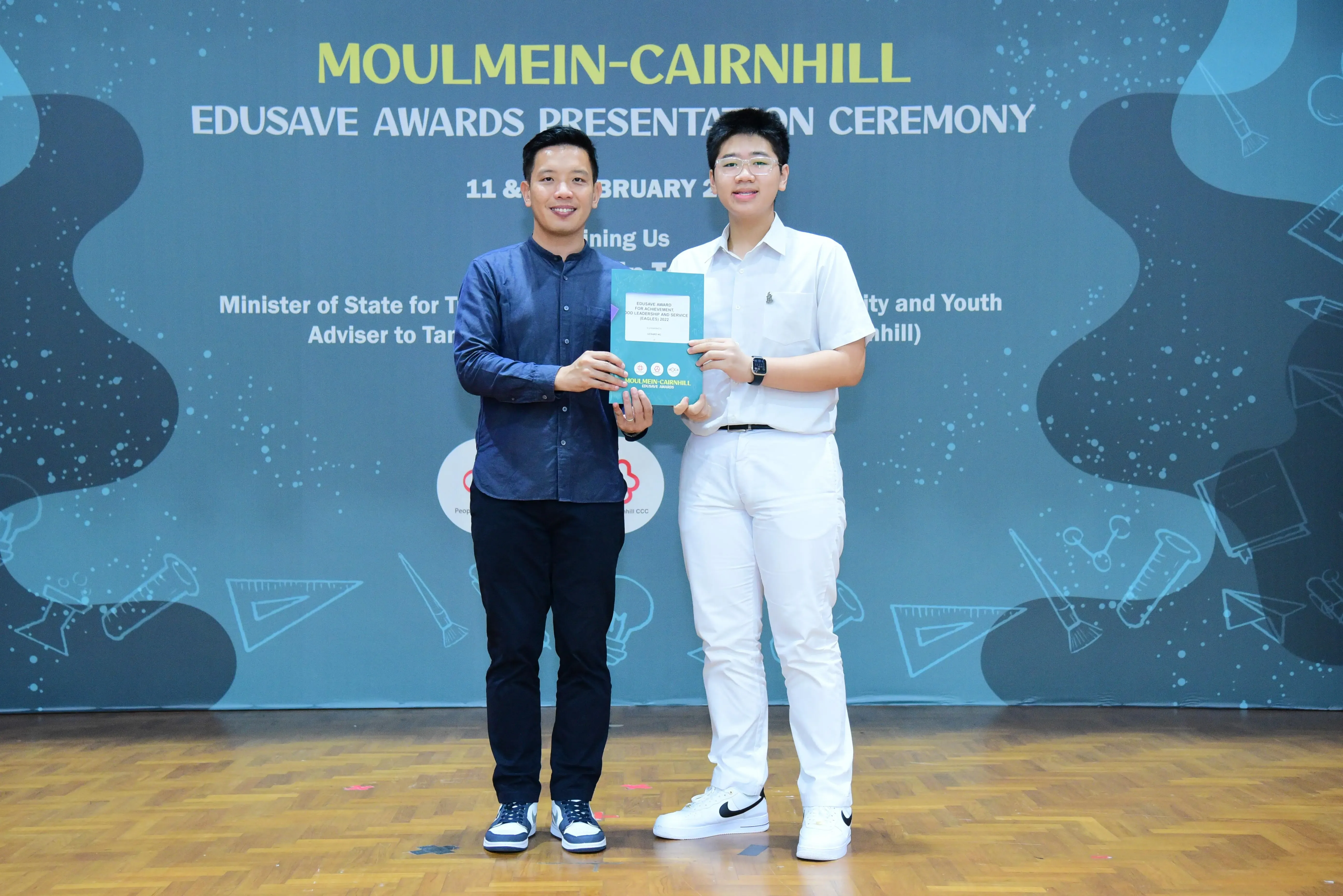 Two men hold an award booklet at the Moulmein-Cairnhill Edusave Awards Presentation Ceremony backdrop.