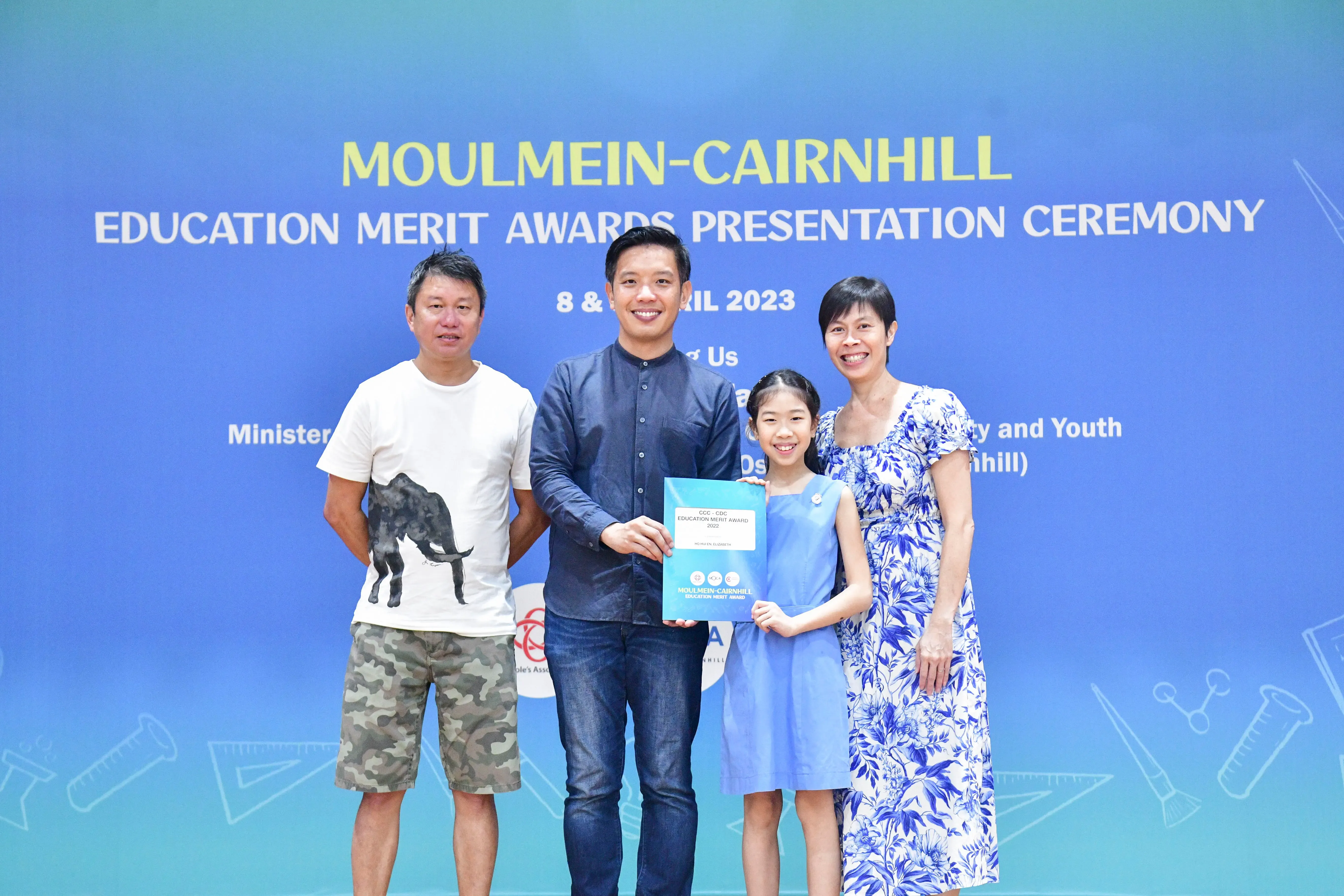 Four people pose before a Moulmein-Cairnhill Education Merit Awards 2023 banner, with a girl holding a certificate.