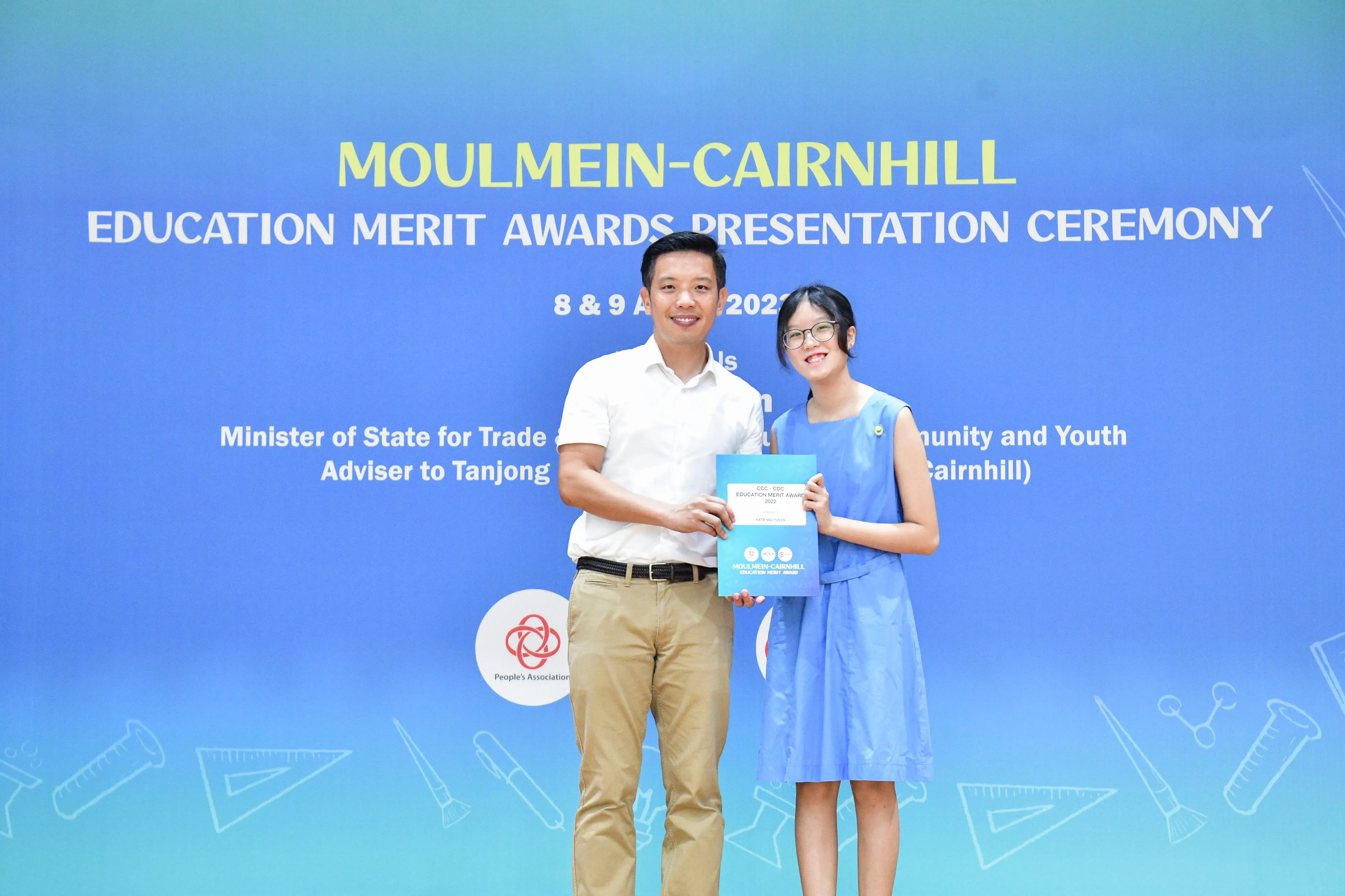 A man and a young girl hold an award certificate in front of a Moulmein-Cairnhill Education Merit Awards banner.