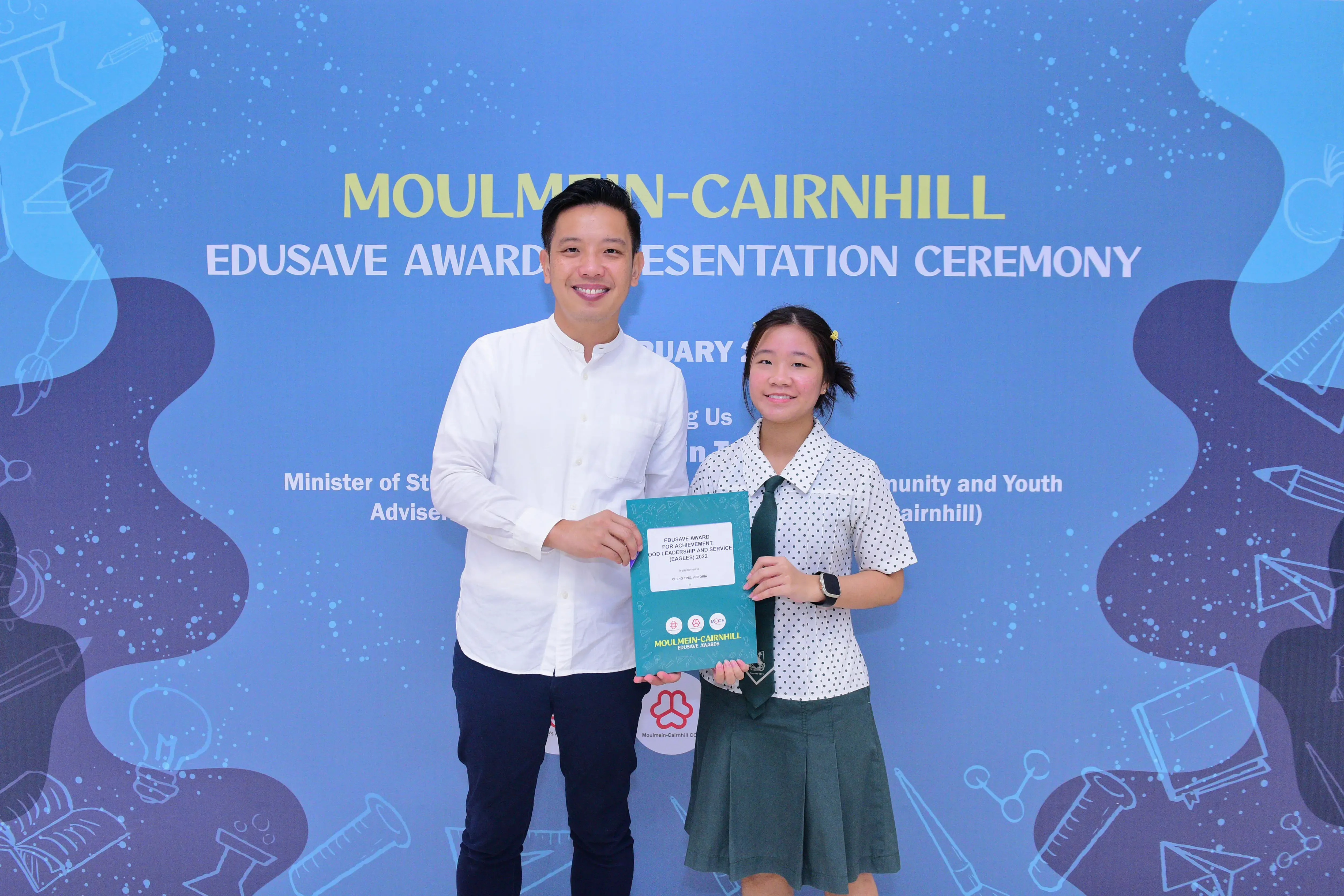 A man and a girl in school uniform hold a certificate at the Moulmein-Cairnhill Edusave Award Presentation Ceremony.