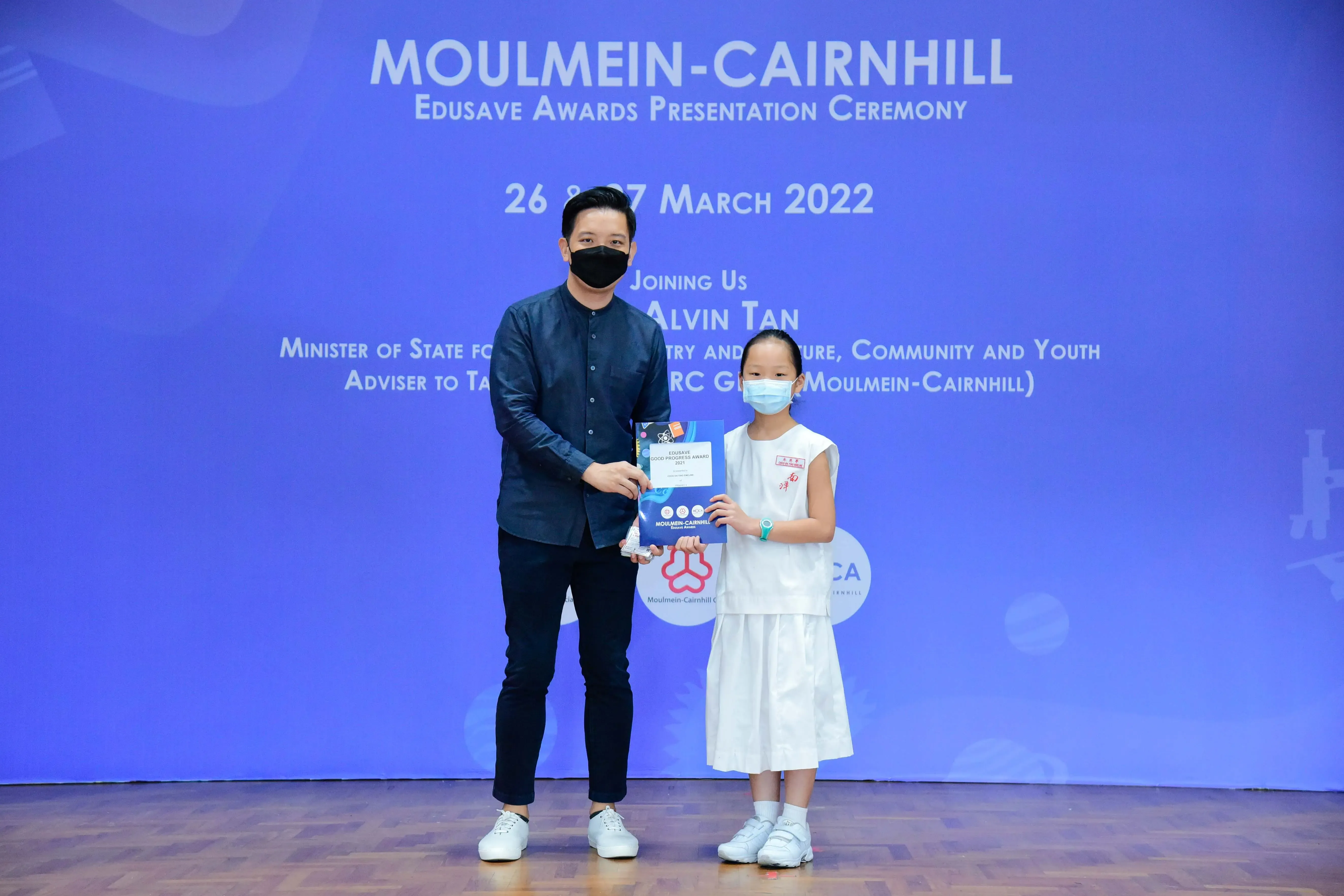 An adult and a young girl in masks hold an award certificate at the Moulmein-Cairnhill Edusave Awards Ceremony, March 2022.