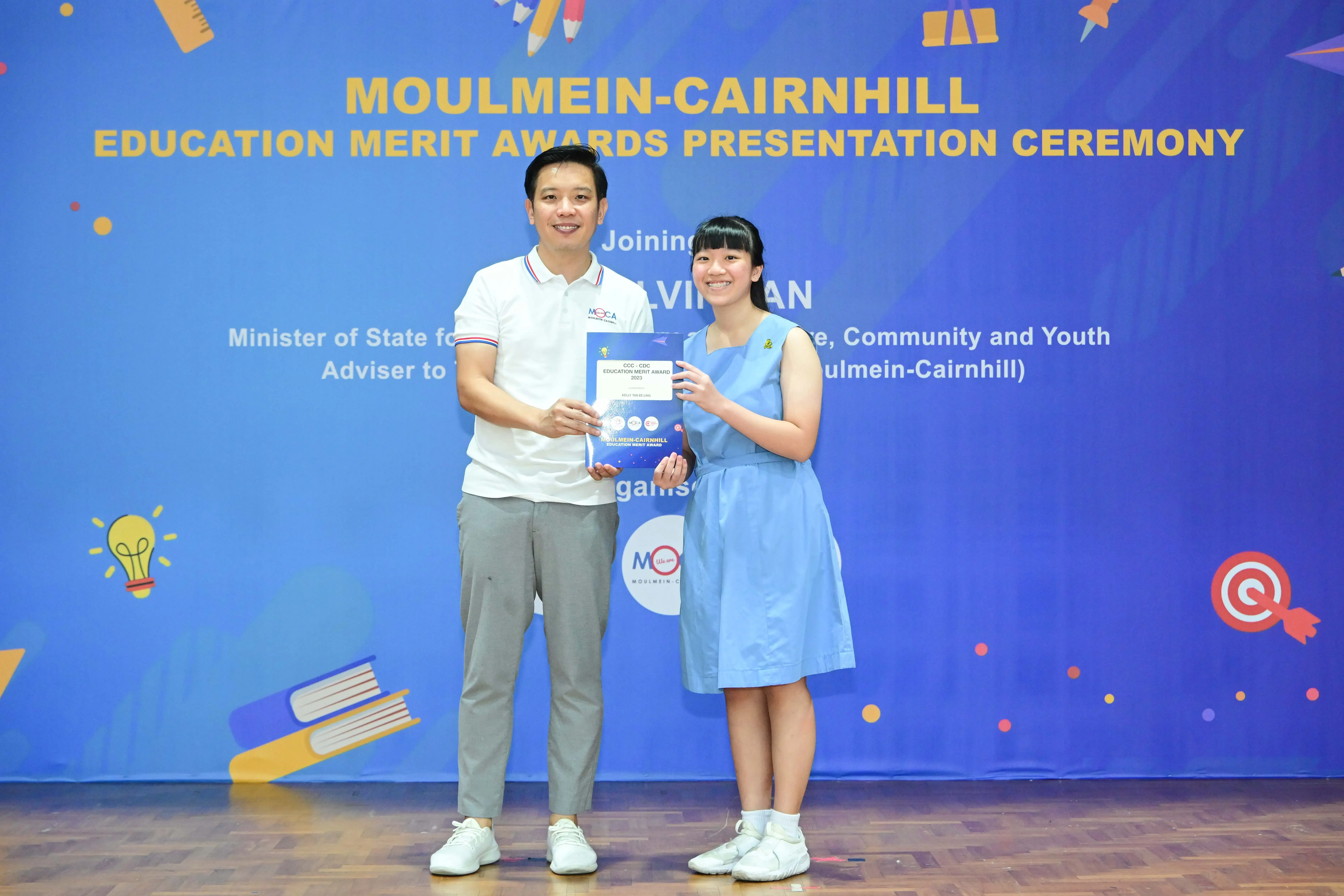 A man and a young girl hold an award certificate together in front of a Moulmein-Cairnhill Education Merit Awards banner.