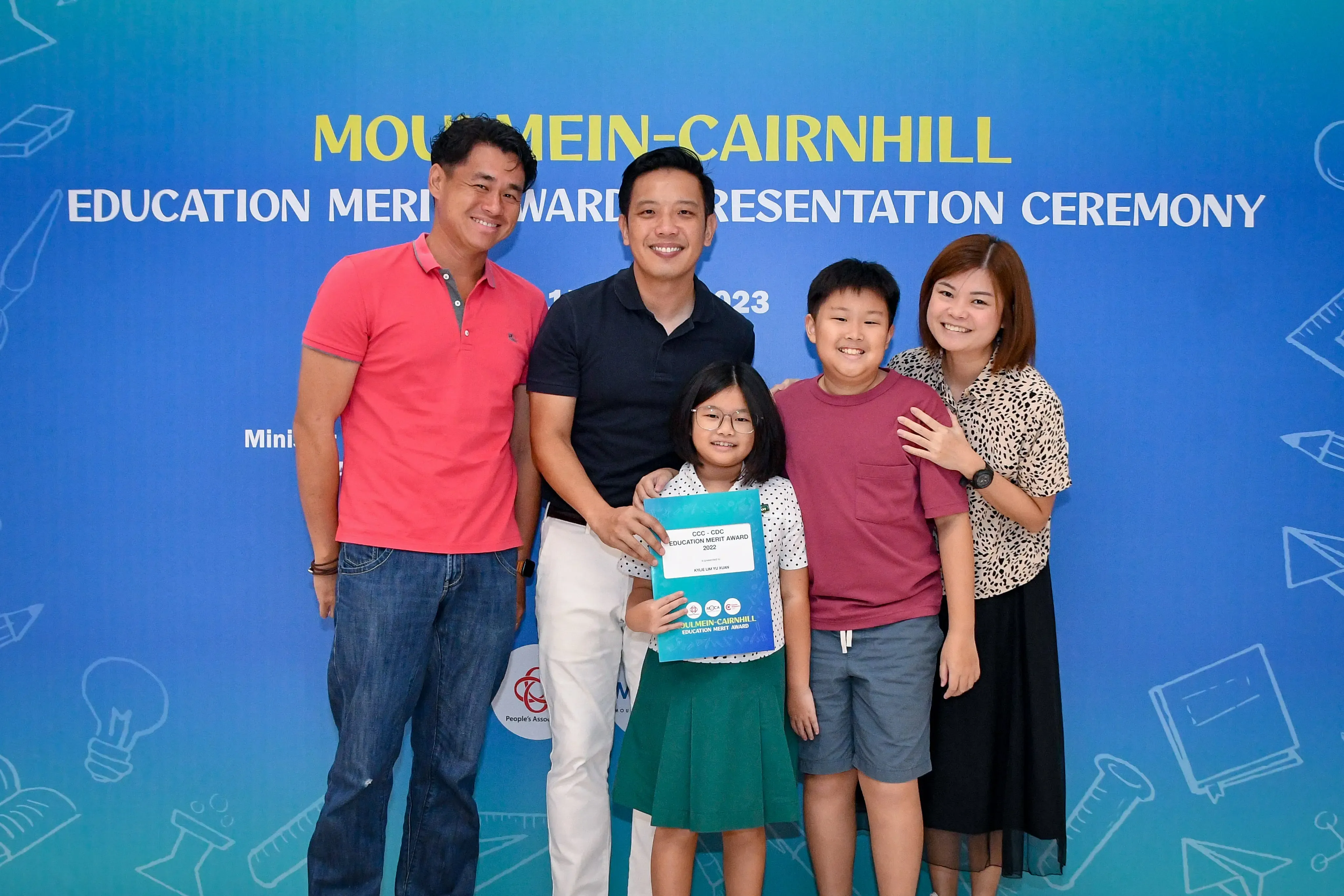 Five people pose smiling before a Moulmein-Cairnhill Education Merit Award Presentation Ceremony banner, girl holds award folder.