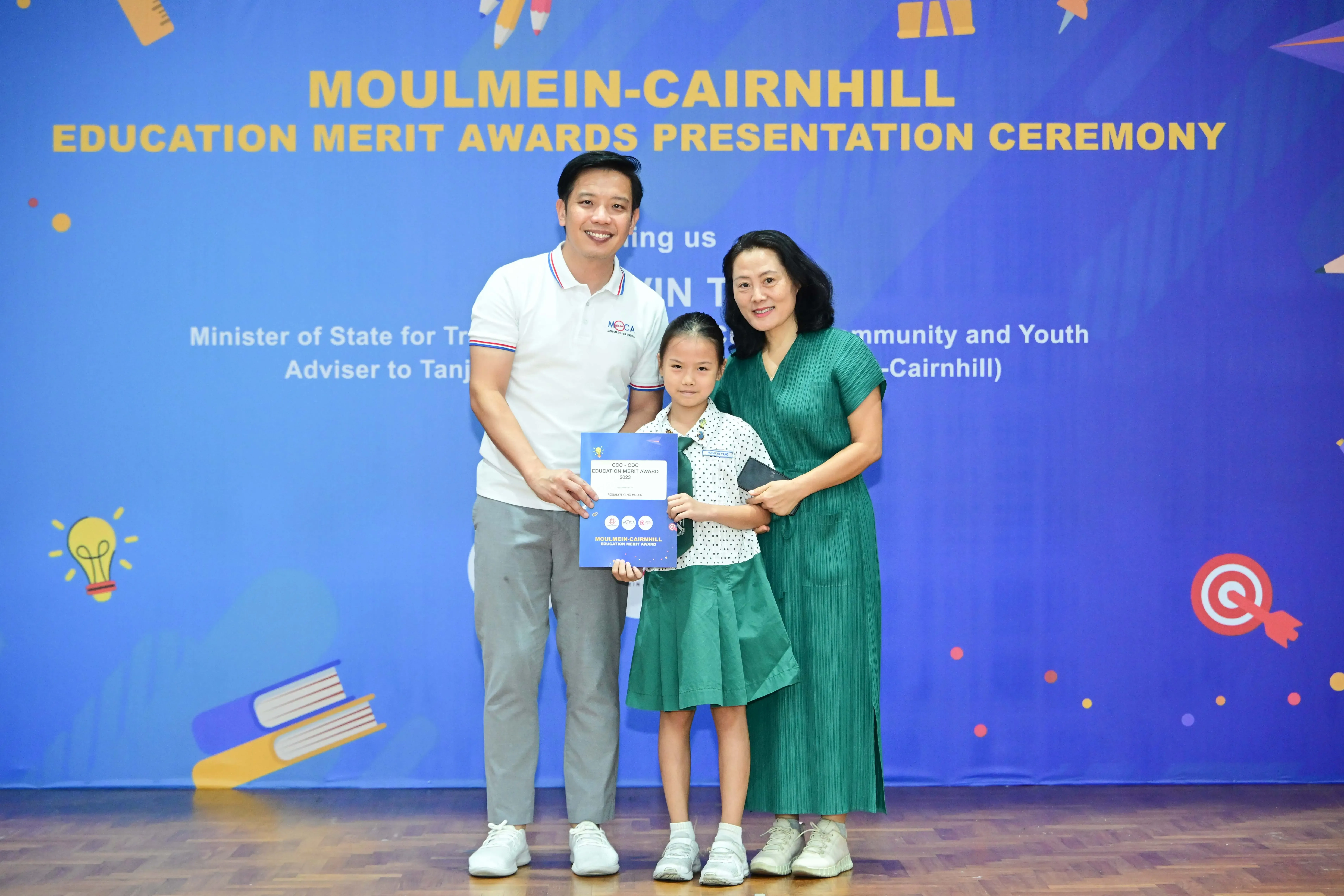 A girl holds a merit award certificate flanked by two adults at the Moulmein-Cairnhill Education Merit Awards Ceremony.
