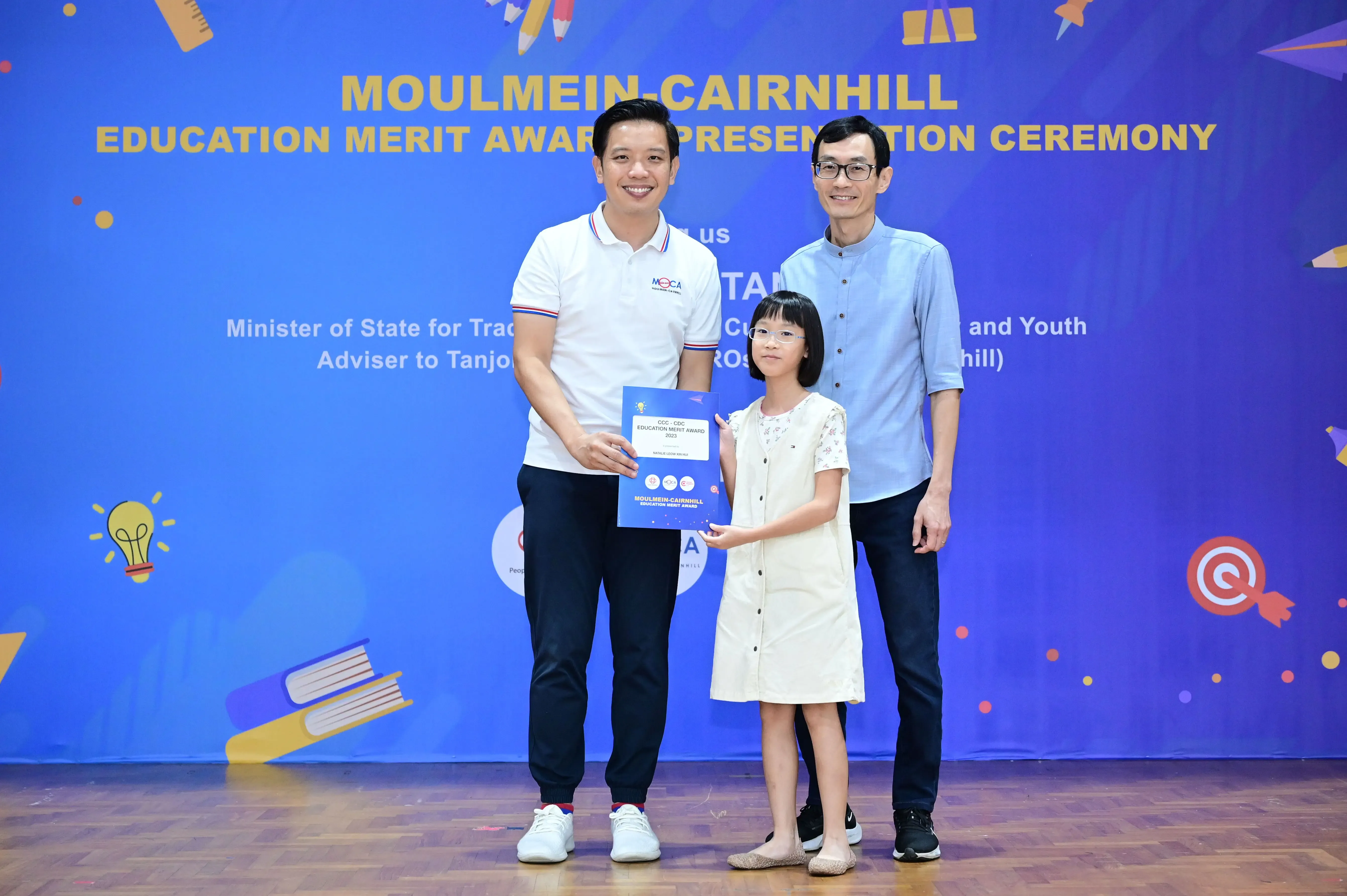 Two men and a young girl hold a certificate at the Moulmein-Cairnhill Education Merit Award Presentation Ceremony.
