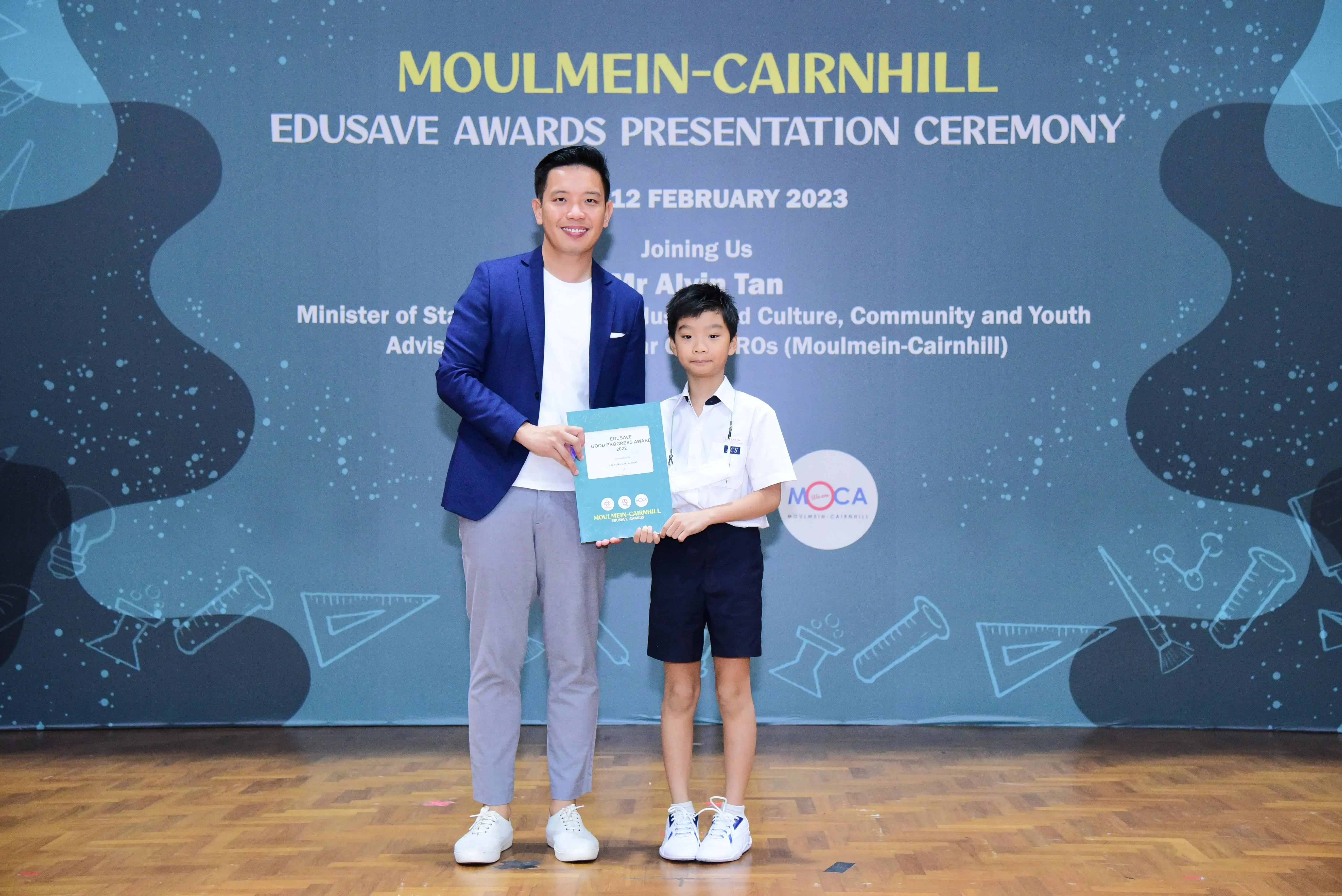 A man in a blue blazer and a young boy in school uniform hold a certificate at the Moulmein-Cairnhill Edusave Awards 2023.