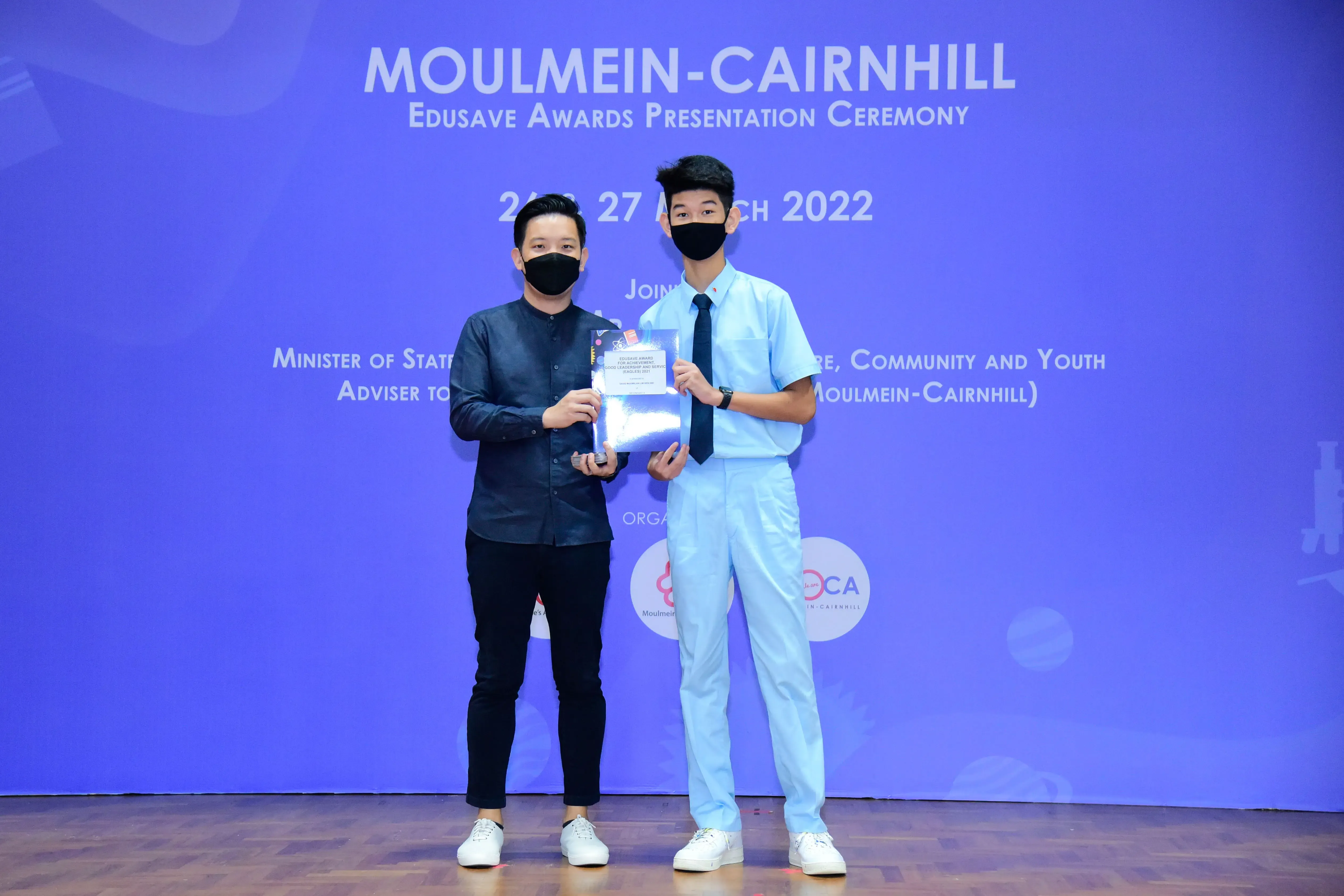 Two masked men hold an award at the Moulmein-Cairnhill Edusave Awards Presentation Ceremony, March 2022.