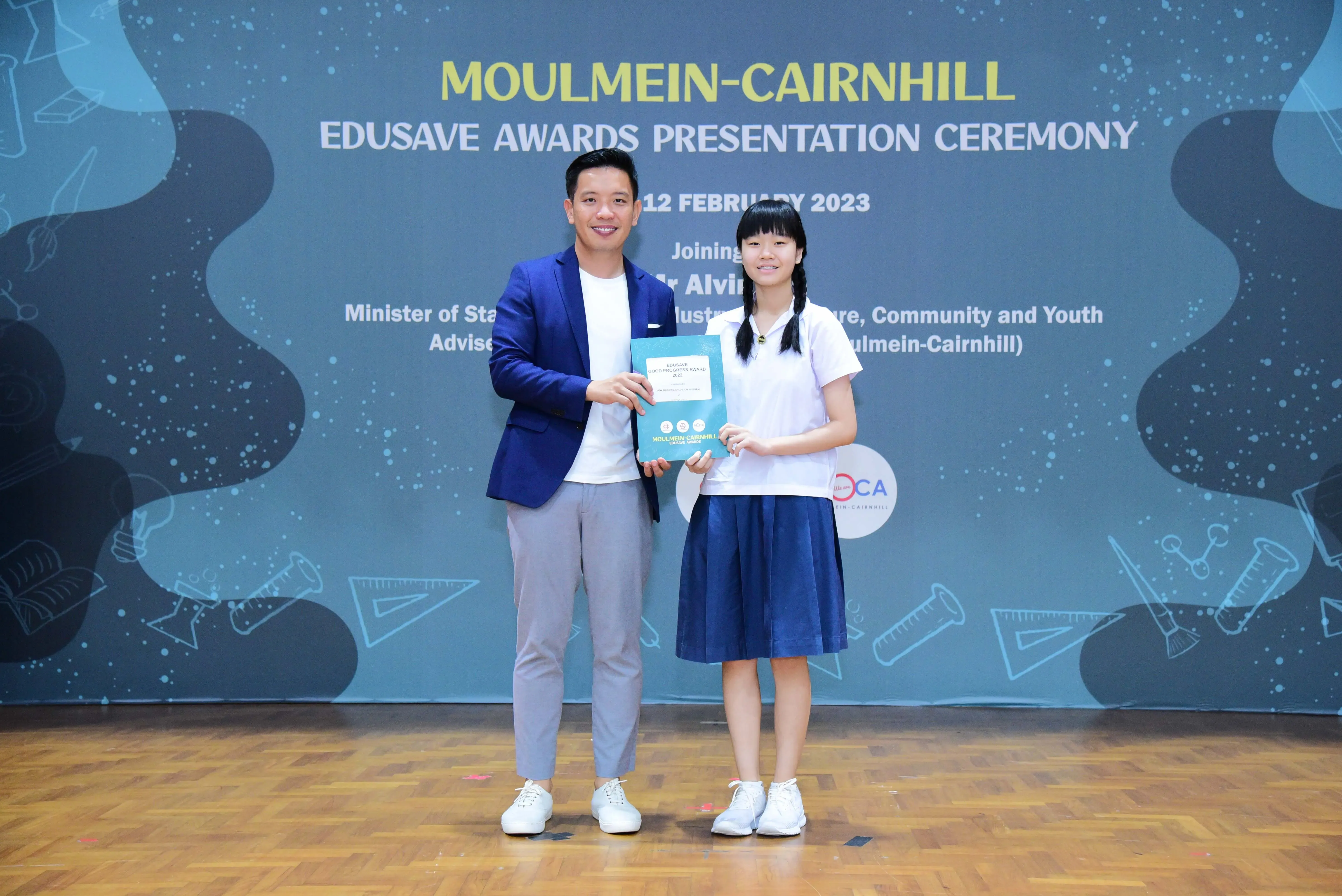 A man in a blue blazer and a young girl in school uniform hold a certificate at the Moulmein-Cairnhill Edusave Awards 2023.