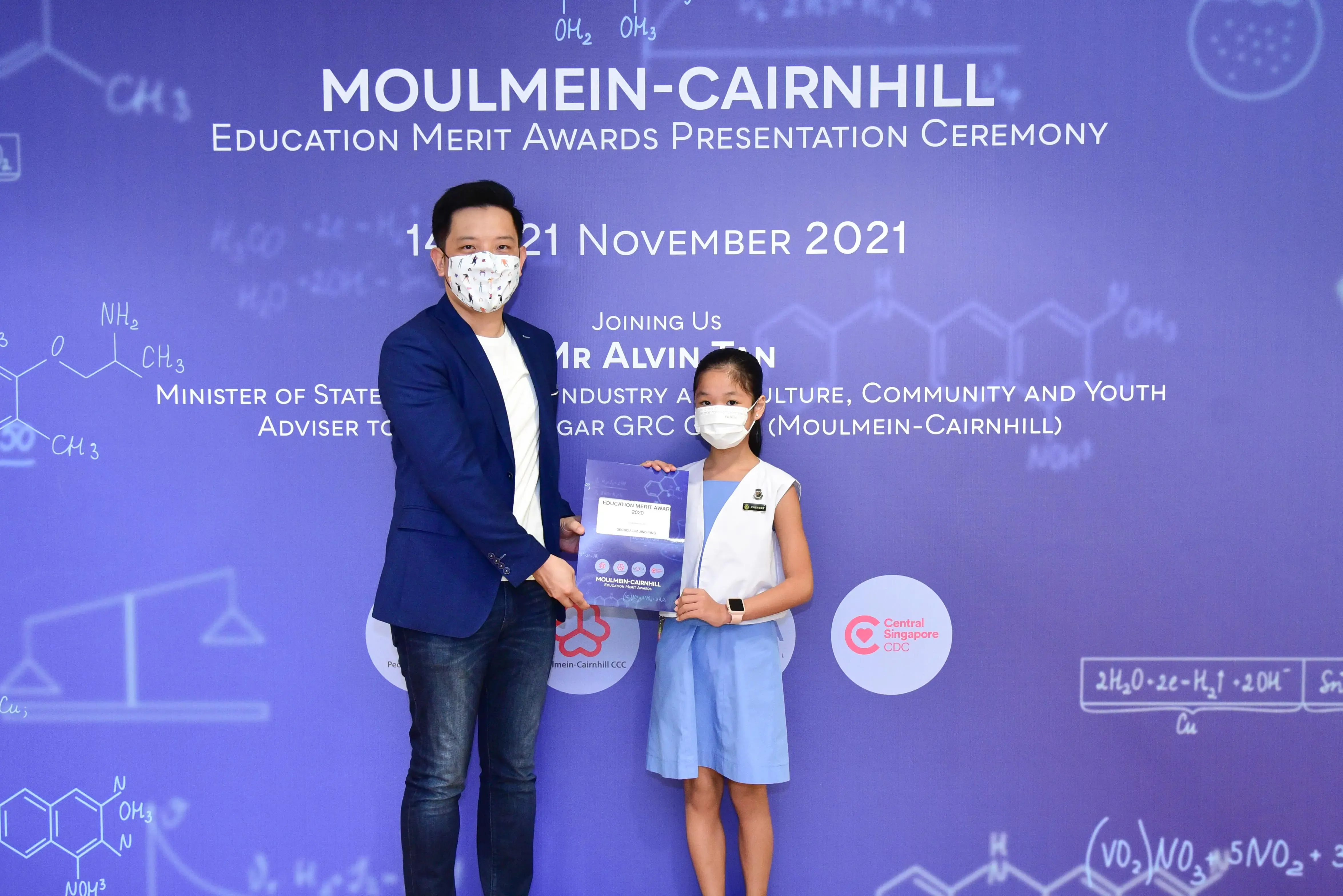 A masked man and young girl hold an award certificate at the Moulmein-Cairnhill Education Merit Awards Ceremony, November 2021.
