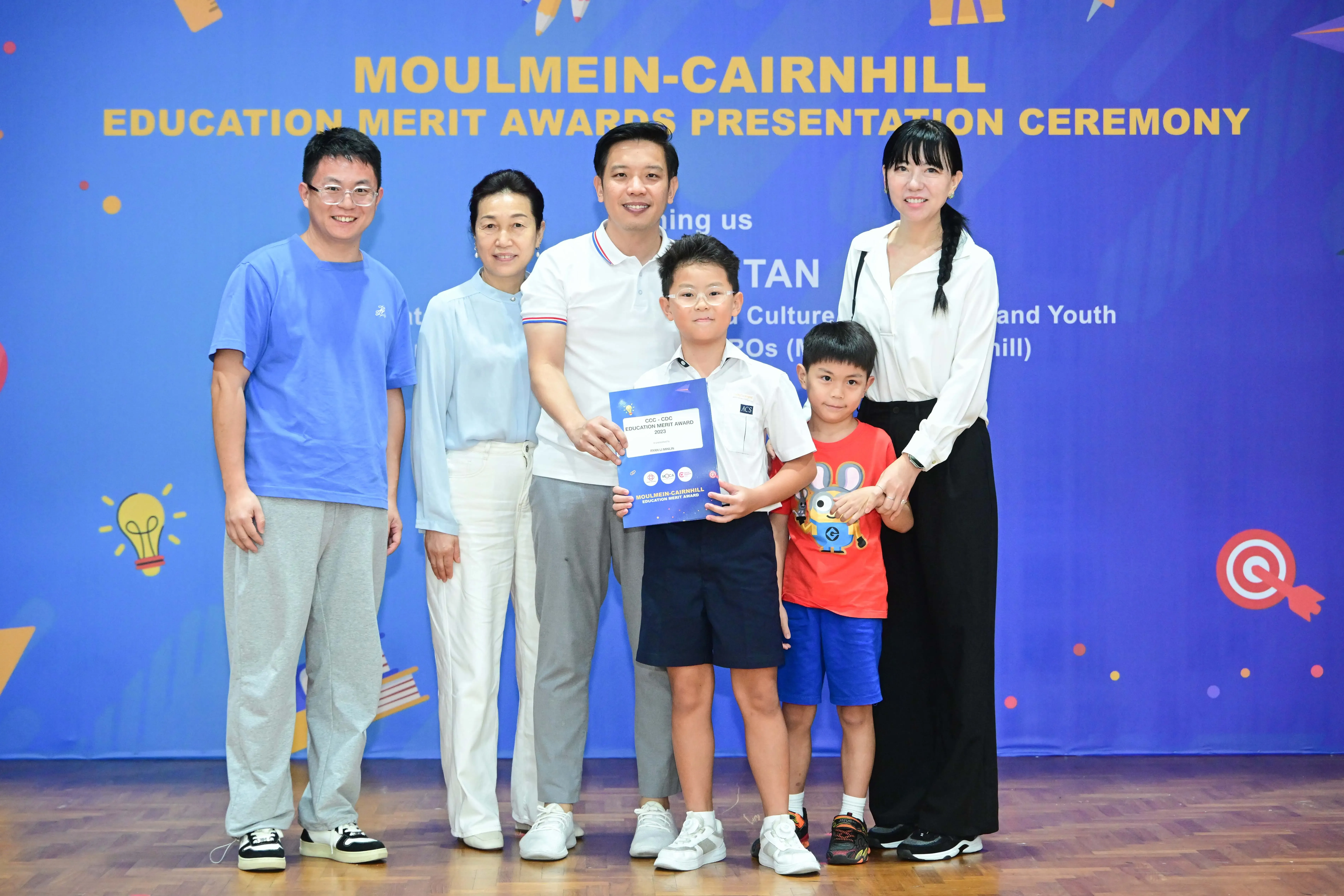 Five adults and two children pose with a certificate at the Moulmein-Cairnhill Education Merit Awards Presentation Ceremony.