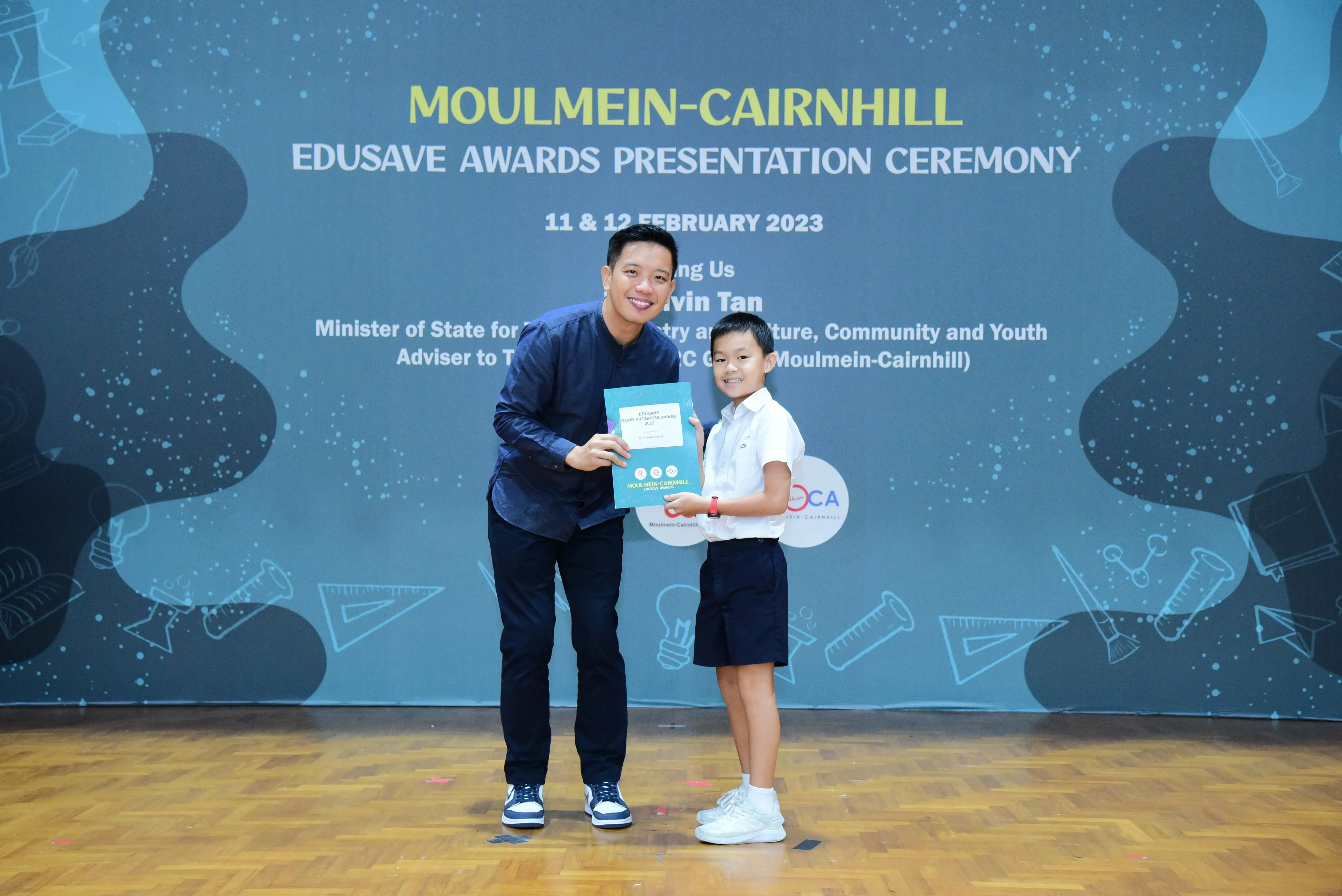 A man and a young boy in school uniform hold a certificate at the Moulmein-Cairnhill Edusave Awards Ceremony 2023.