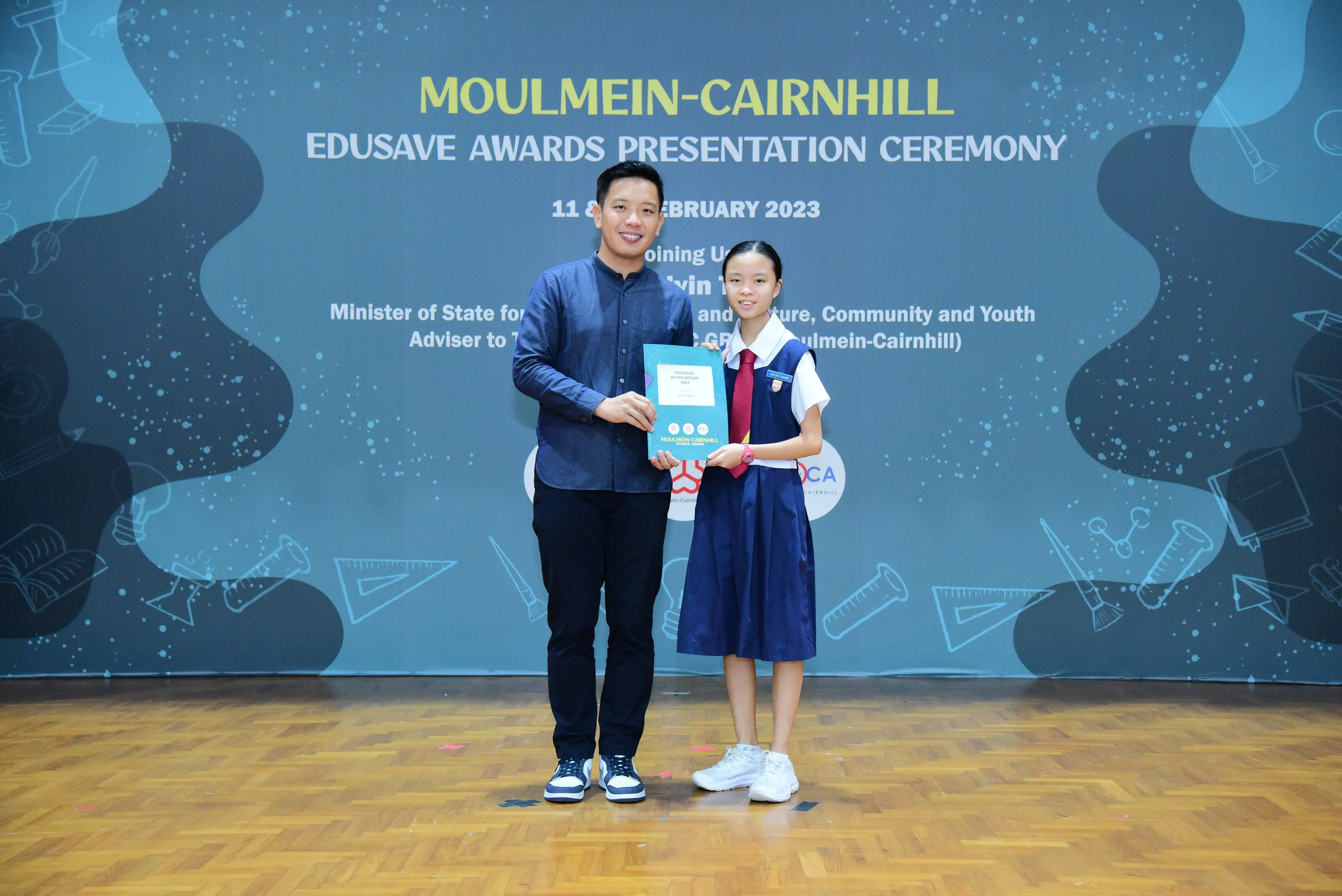 A man and a young girl in school uniform hold a certificate at the Moulmein-Cairnhill Edusave Awards Ceremony 2023.