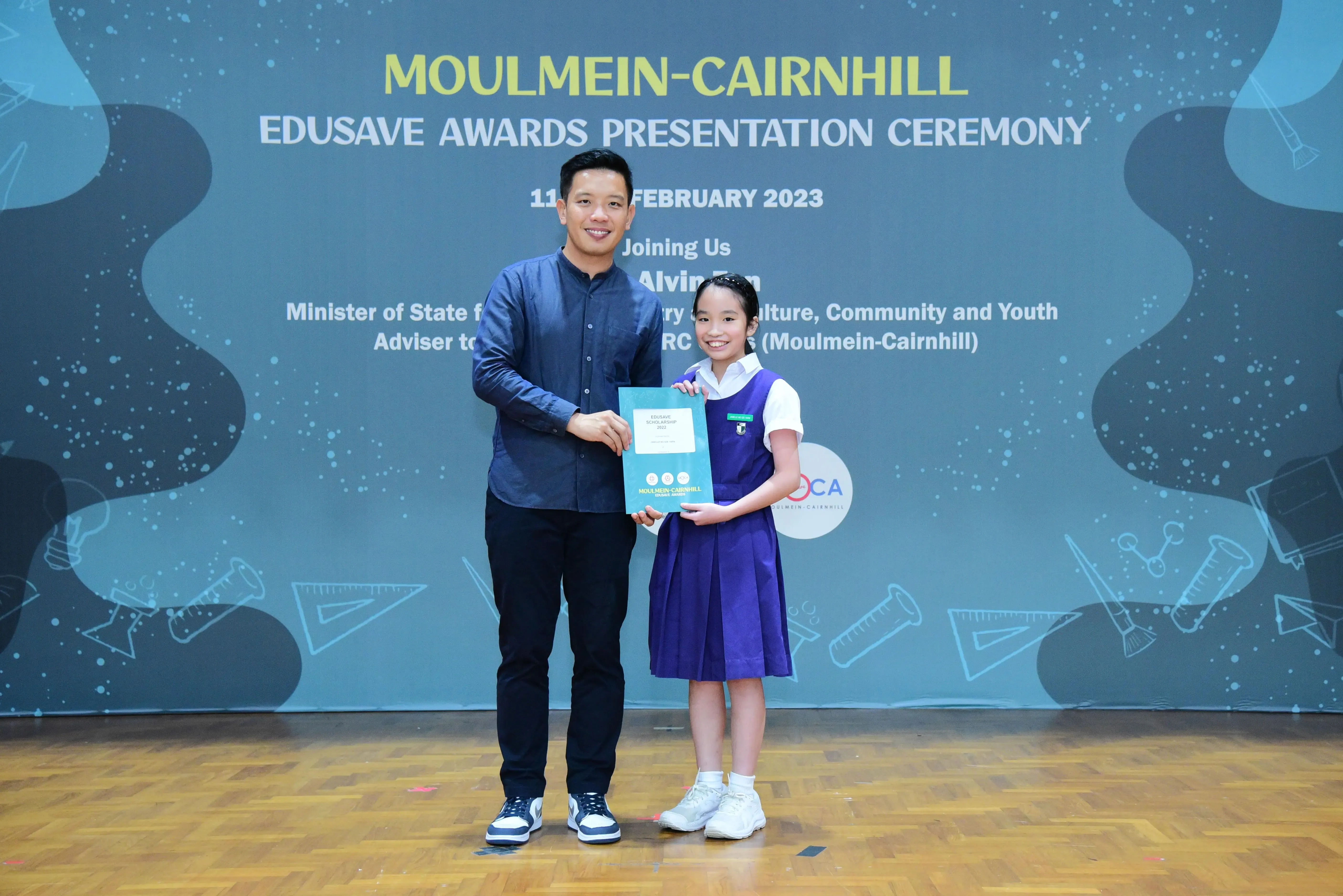 A man and a young girl in school uniform hold a certificate at the Moulmein-Cairnhill Edusave Awards Ceremony 2023.