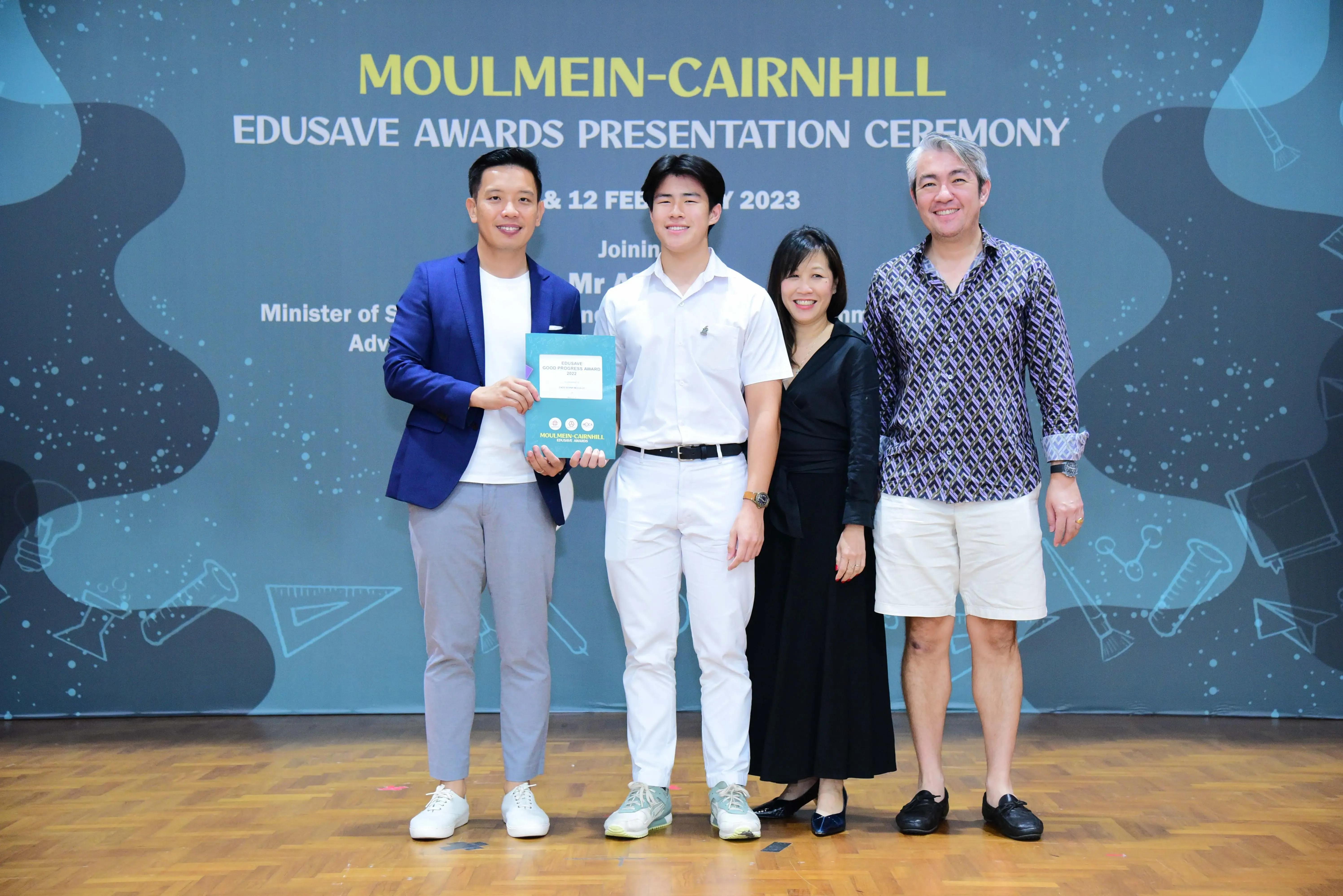 Four people smile on stage at the Moulmein-Cairnhill Edusave Awards Presentation Ceremony 2023, holding an award booklet.